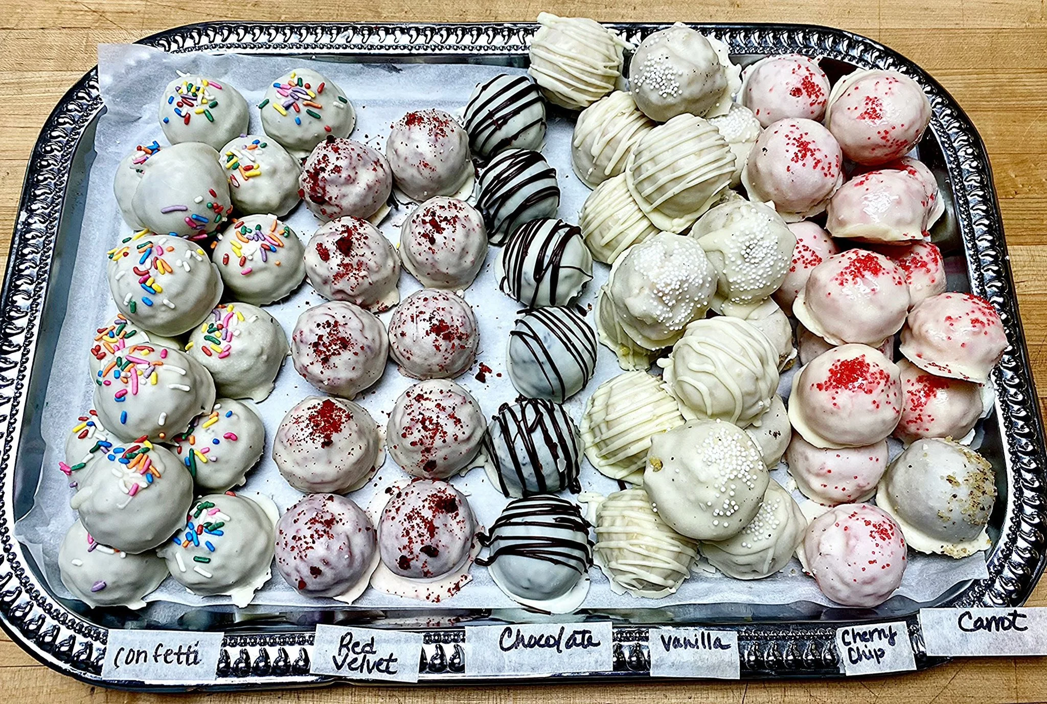 A large silver tray holding an assortment of decorated cake balls. The cake balls are coated in white, black, or pink icing with various toppings like sprinkles and colored sugar. The tray is lined with white parchment paper and has small handwritten labels describing flavors such as confetti, red velvet, chocolate, vanilla, cherry chip, and carrot.