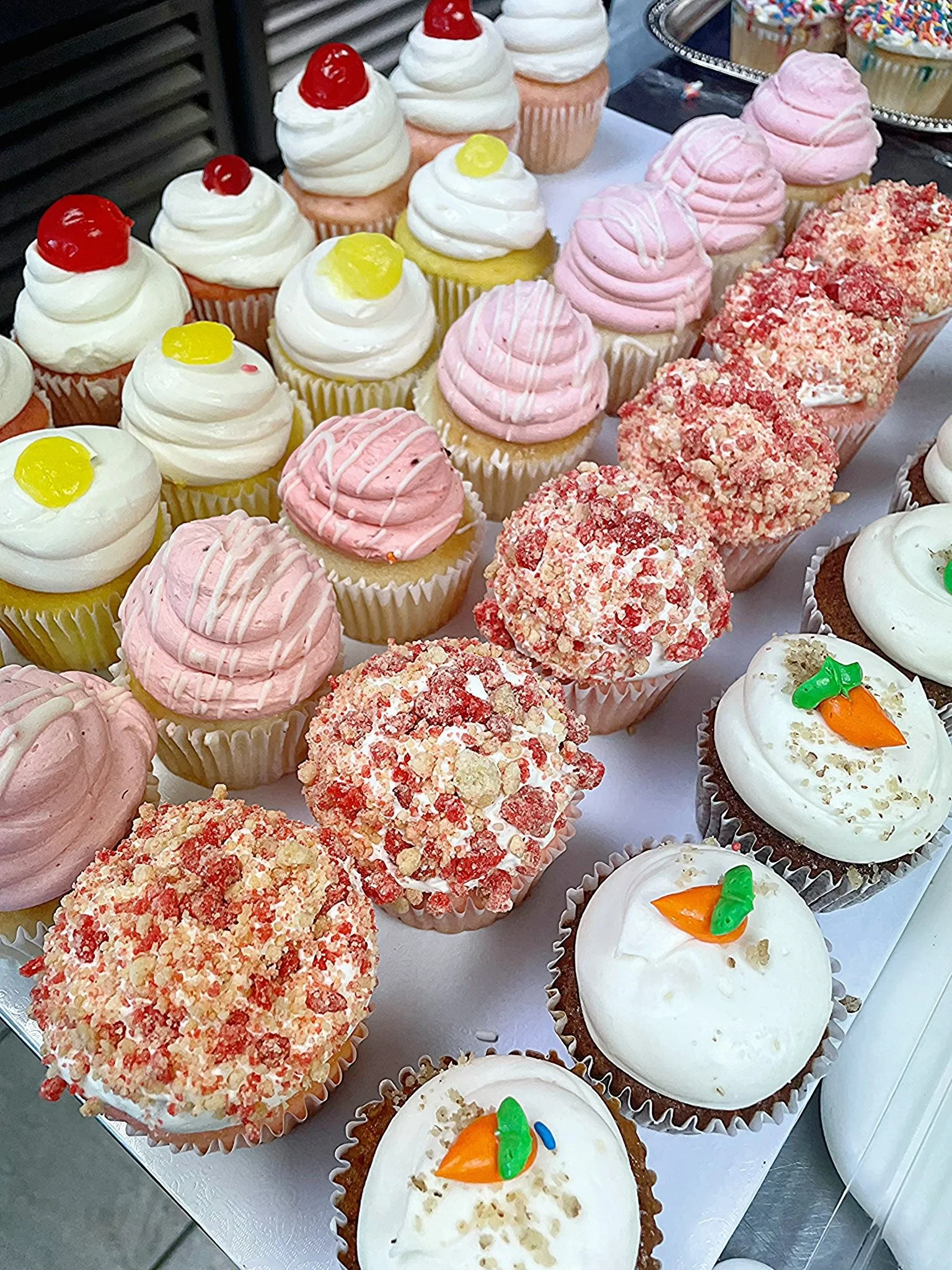 Assorted cupcakes decorated with various toppings, including cherries, yellow candies, pink frosting, white frosting with colorful sprinkles, and some with crumbly red topping, displayed on a table.