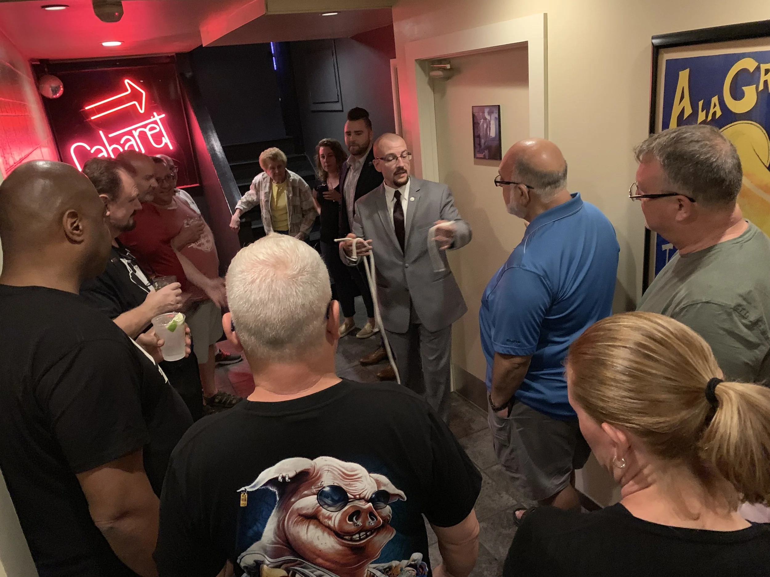 A group of people gathered around a man in a gray suit who is performing a magic trick with a rope in a dimly lit bar or restaurant. The background features a neon sign that says "Cabaret" and a colorful framed poster.