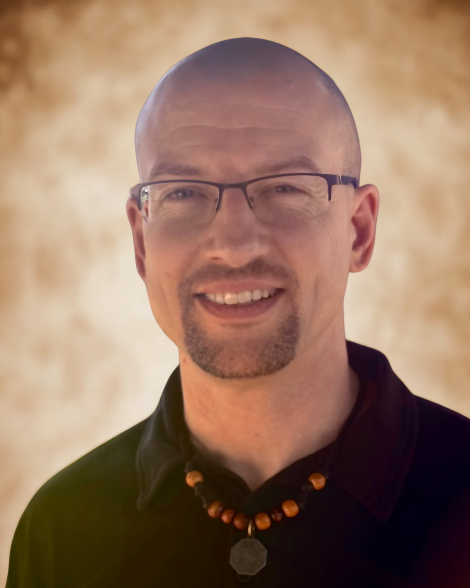 A smiling man with glasses, a goatee, and a shaved head, wearing a black shirt and a beaded necklace with a pendant, standing in front of a blurred, warm-toned background.