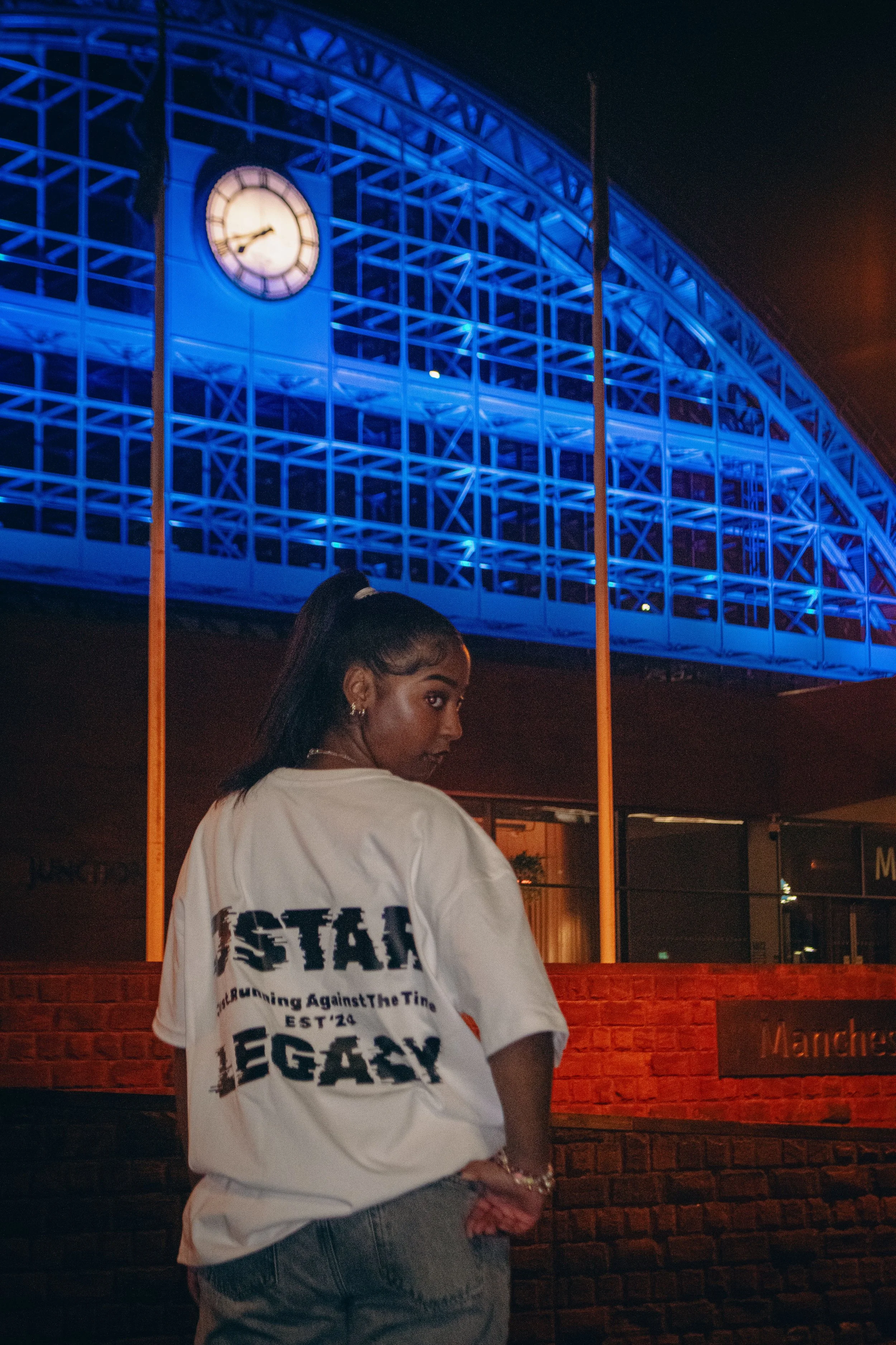 A young woman with dark hair in a high ponytail and hoop earrings, wearing a white T-shirt with text on the back, standing in front of a brick wall with a sign that says 'Manchester.' She is looking over her shoulder at the camera, with her left hand