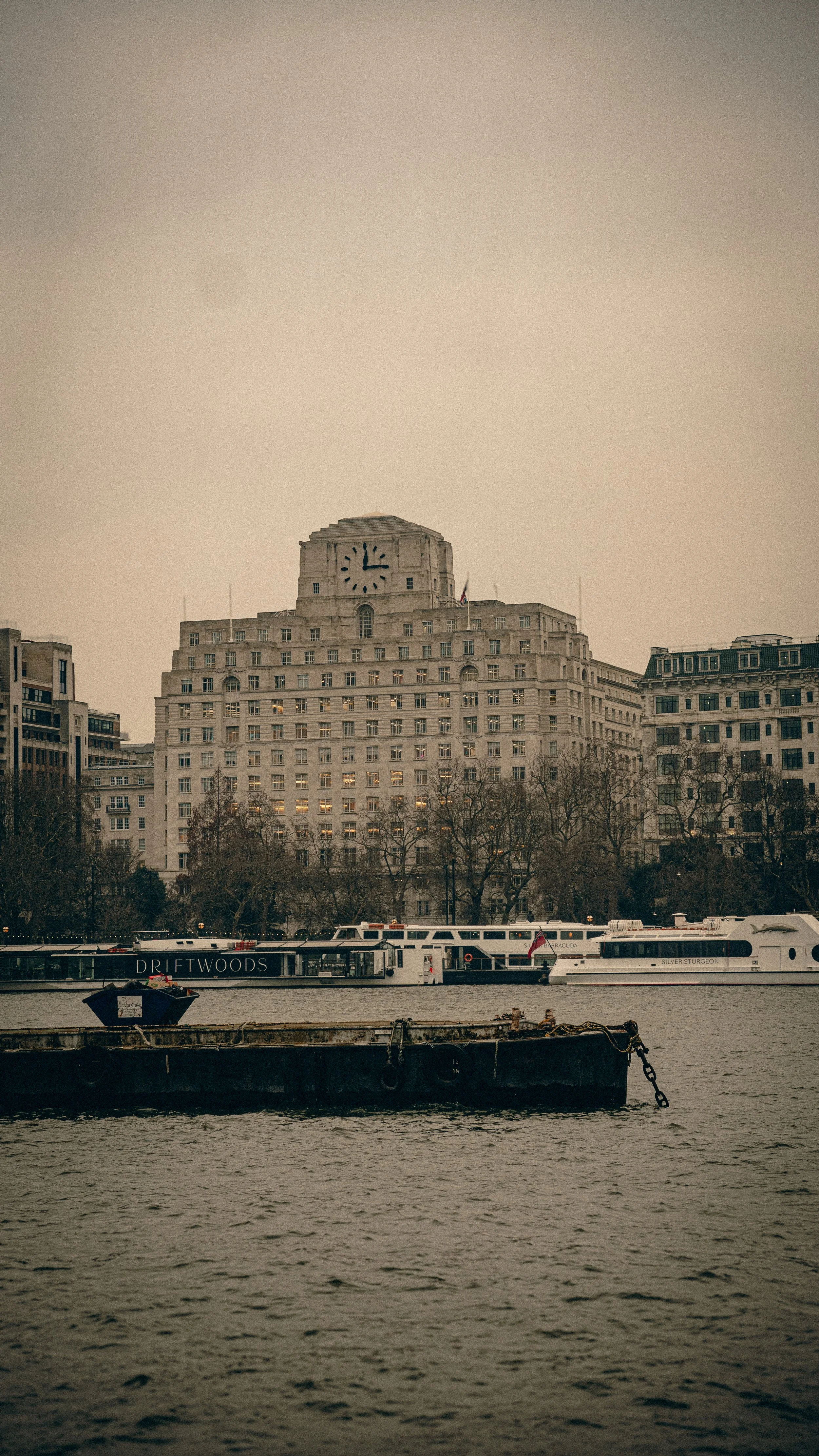 A cityscape with a large historic building near a river, featuring a clock tower, office windows, and boats on the water.