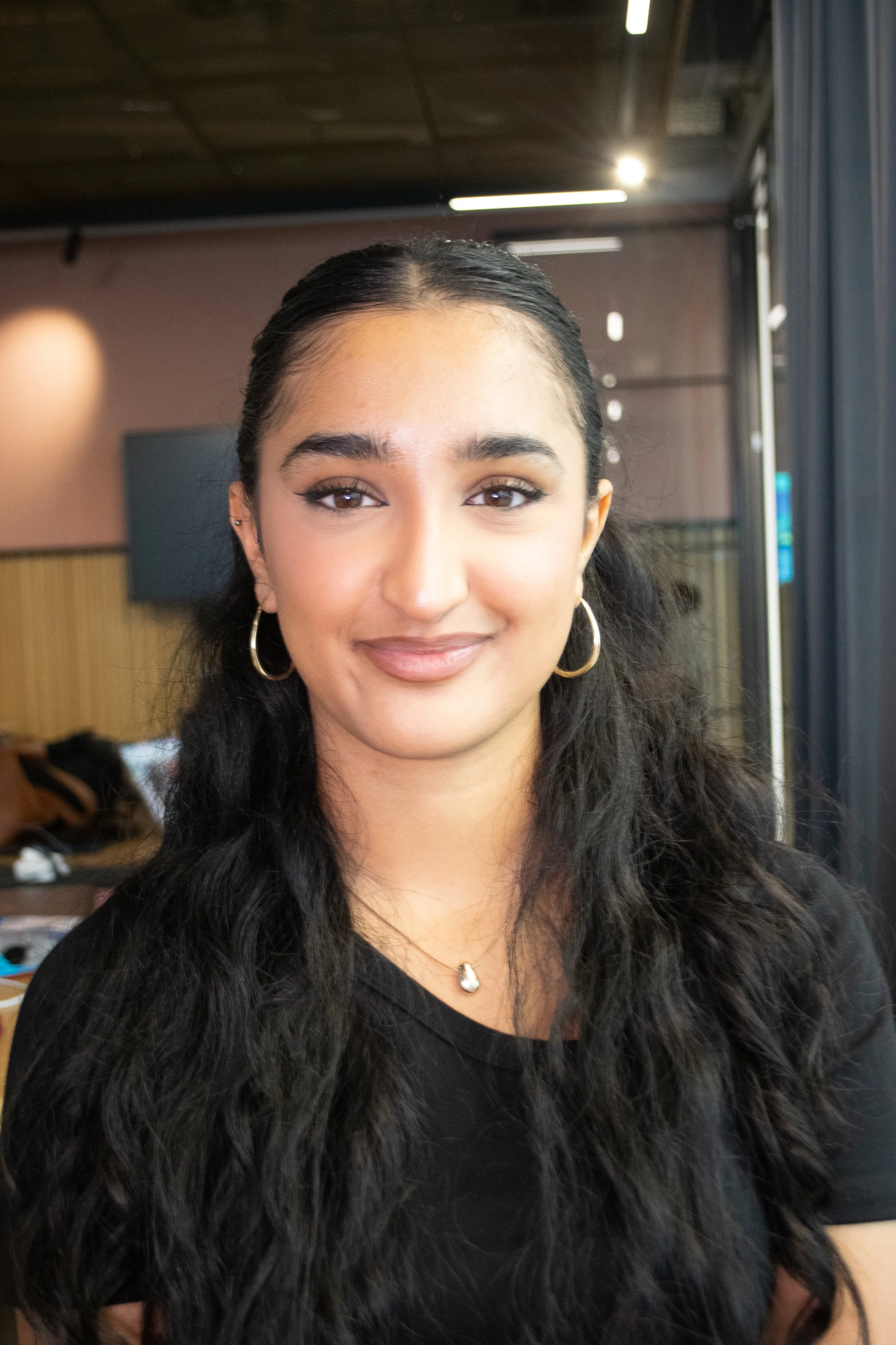 A young woman with long, wavy black hair, wearing hoop earrings and a black top, smiling and standing indoors near a window.