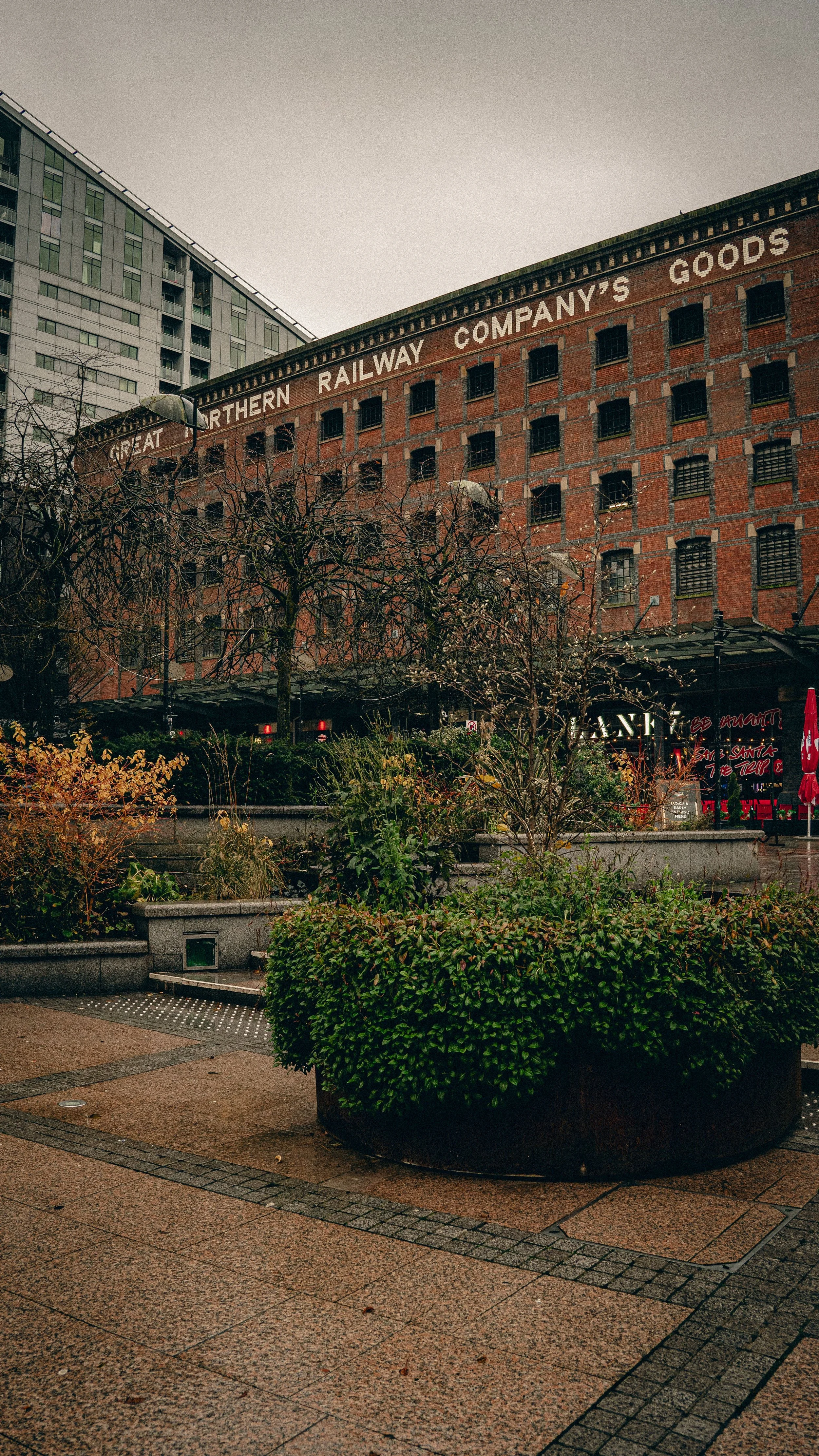 A city street scene featuring a large brick building with the sign 'Great Northern Railway Company's Goods' and leafless trees in an urban park area with benches and shrubs.