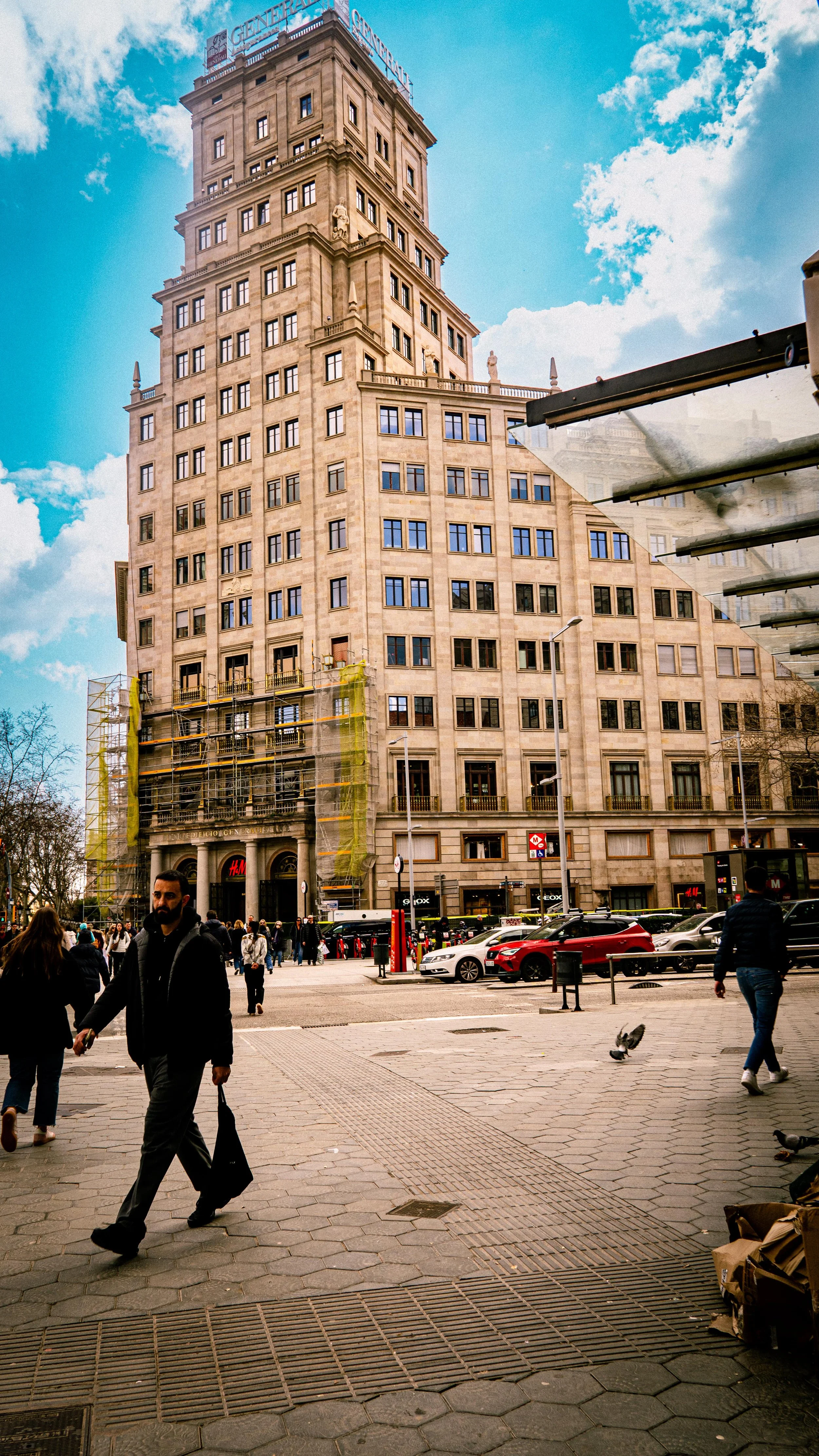 A tall, beige building with a modern architectural design, situated in an urban area with people walking on the sidewalk and cars parked nearby, under a partly cloudy sky.