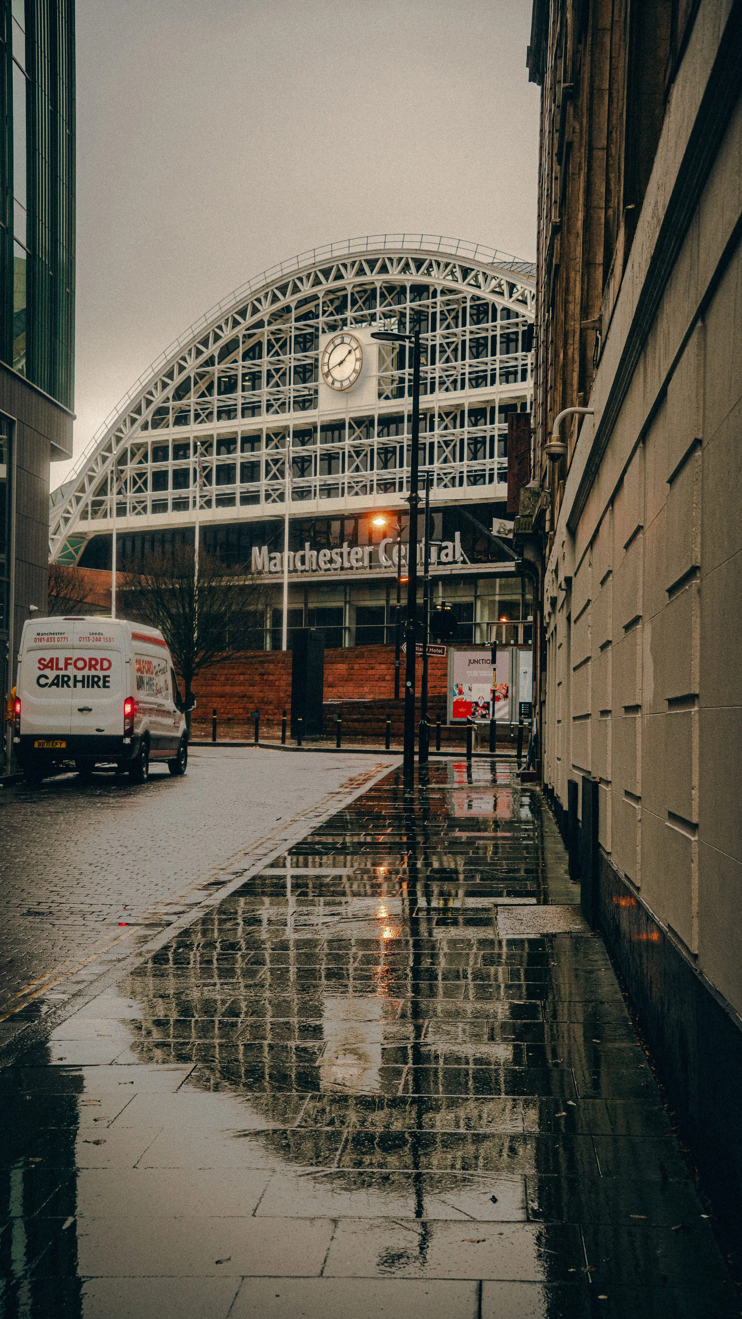 Street view of Manchester Central, with a wet sidewalk reflecting the building and a van parked nearby.