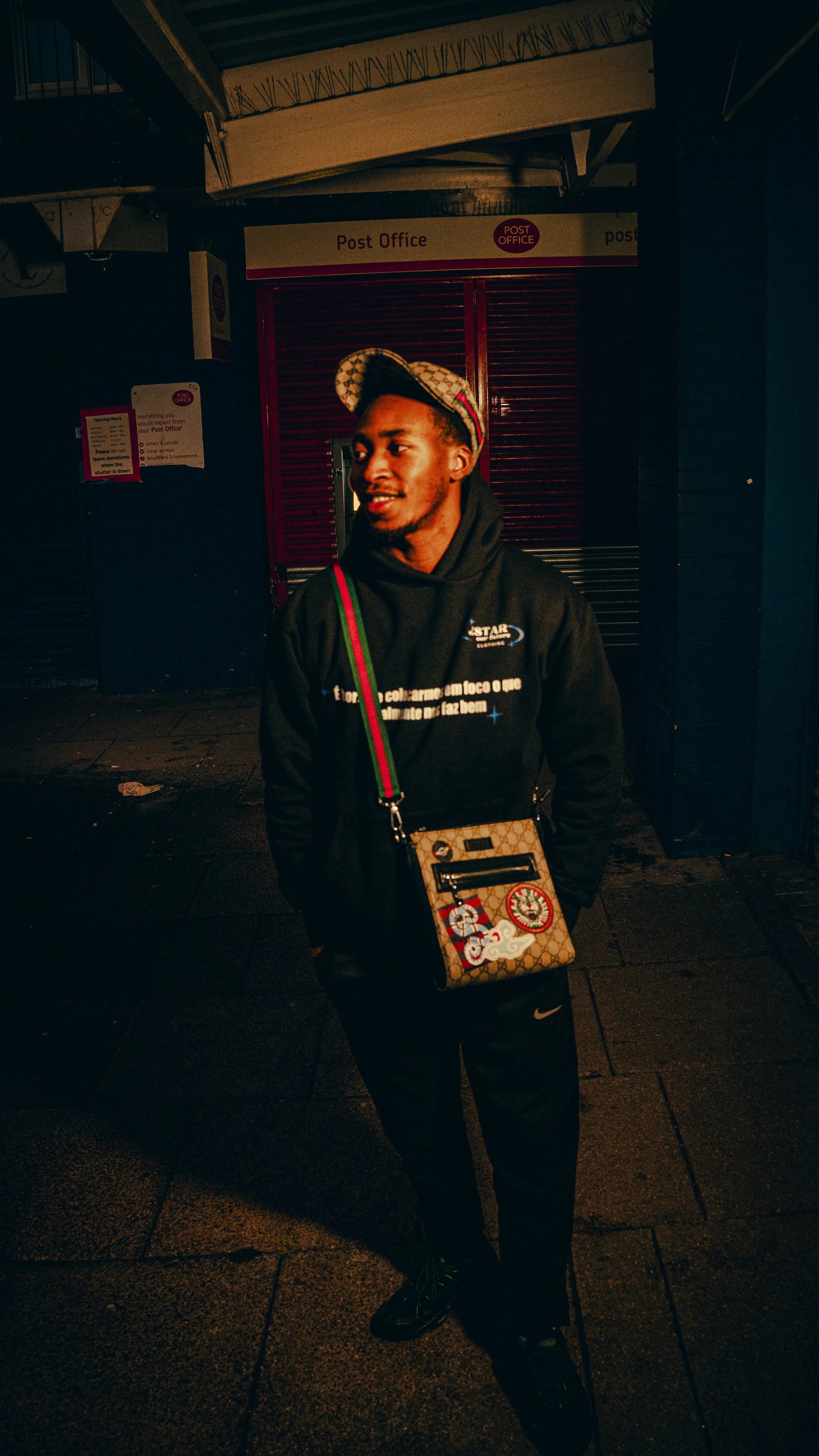 A young man standing outside at night in front of a closed post office, wearing a hoodie, baseball cap, and carrying a patterned bag with patches and patches.