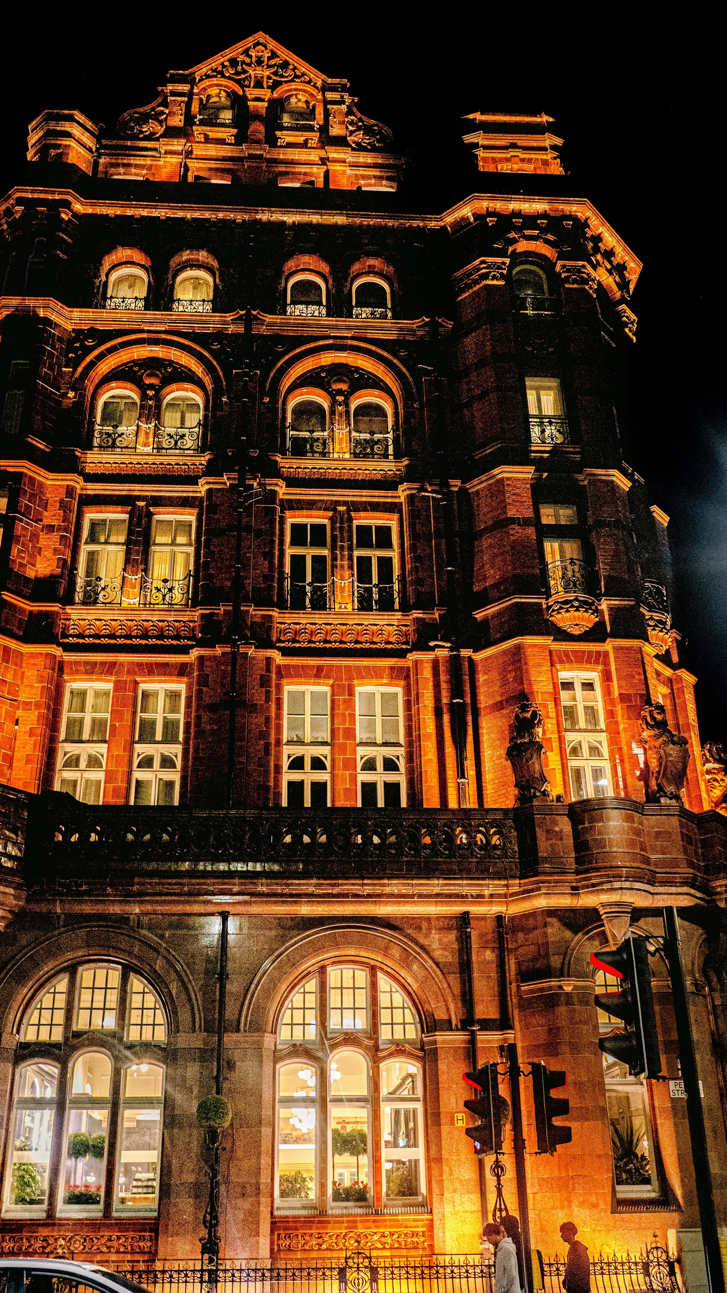 A tall, historic brick building illuminated at night with decorative details, arched windows, and a small balcony. Pedestrians and traffic lights are visible in the foreground.