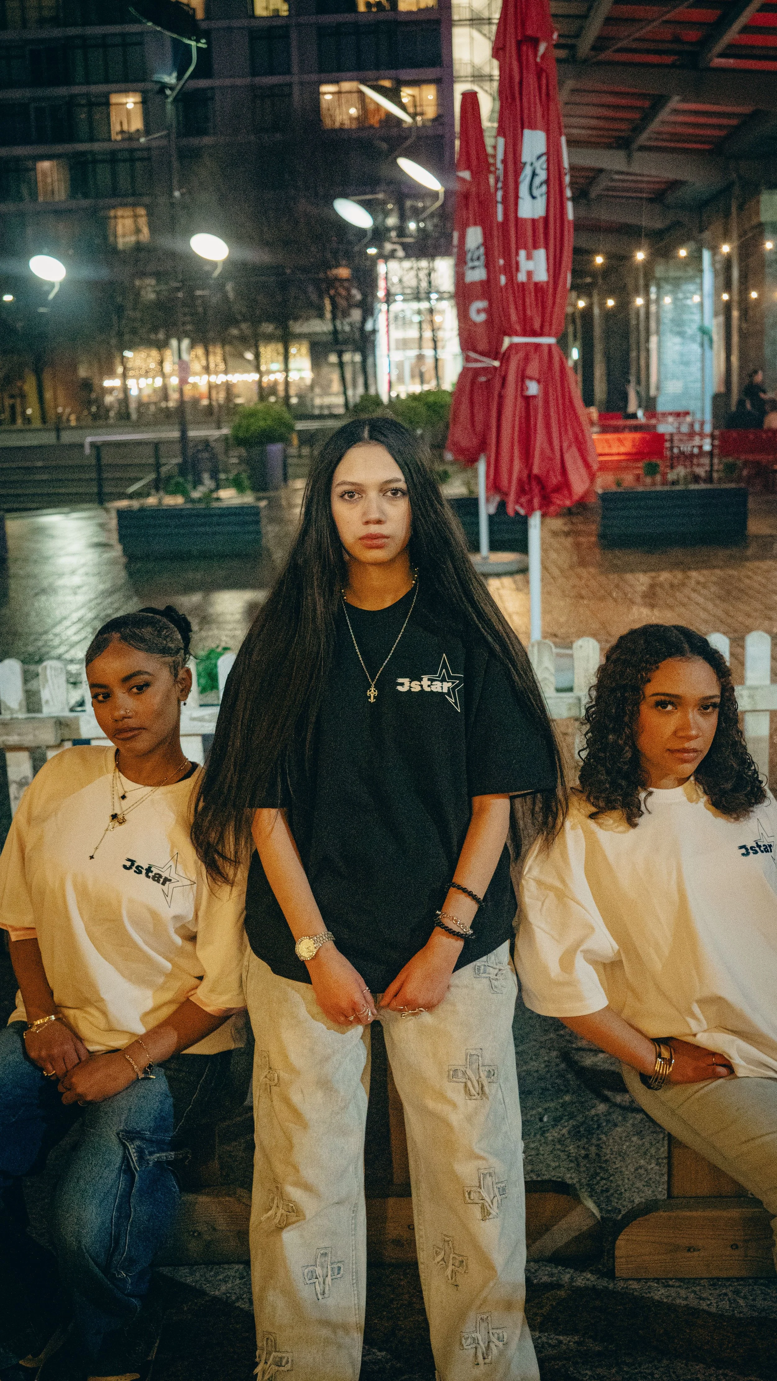 Three women sitting outdoors at night, two wearing off-white t-shirts with