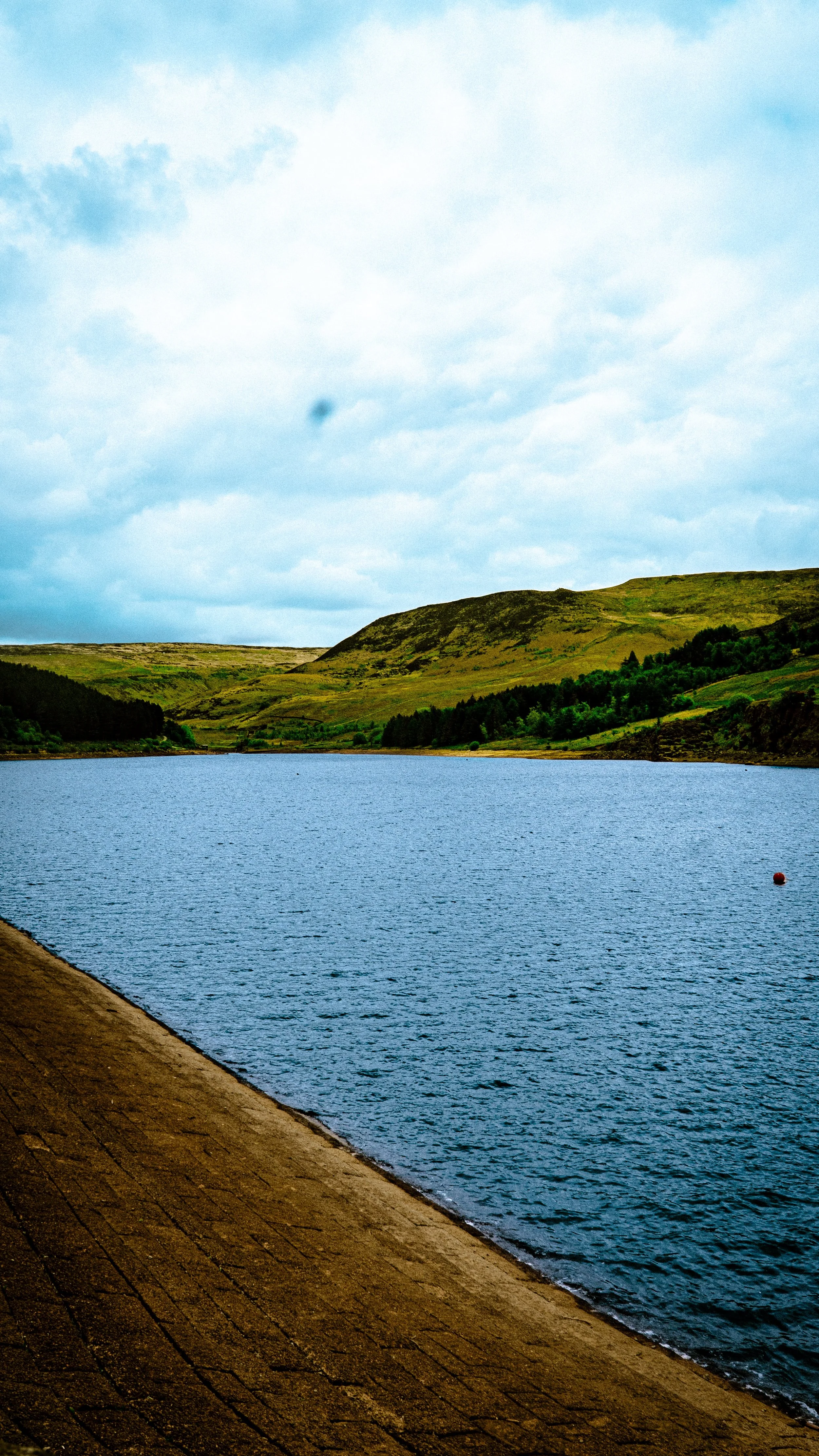 A calm lake with a grassy hillside in the background, partly cloudy sky, and a brick pathway along the water's edge.