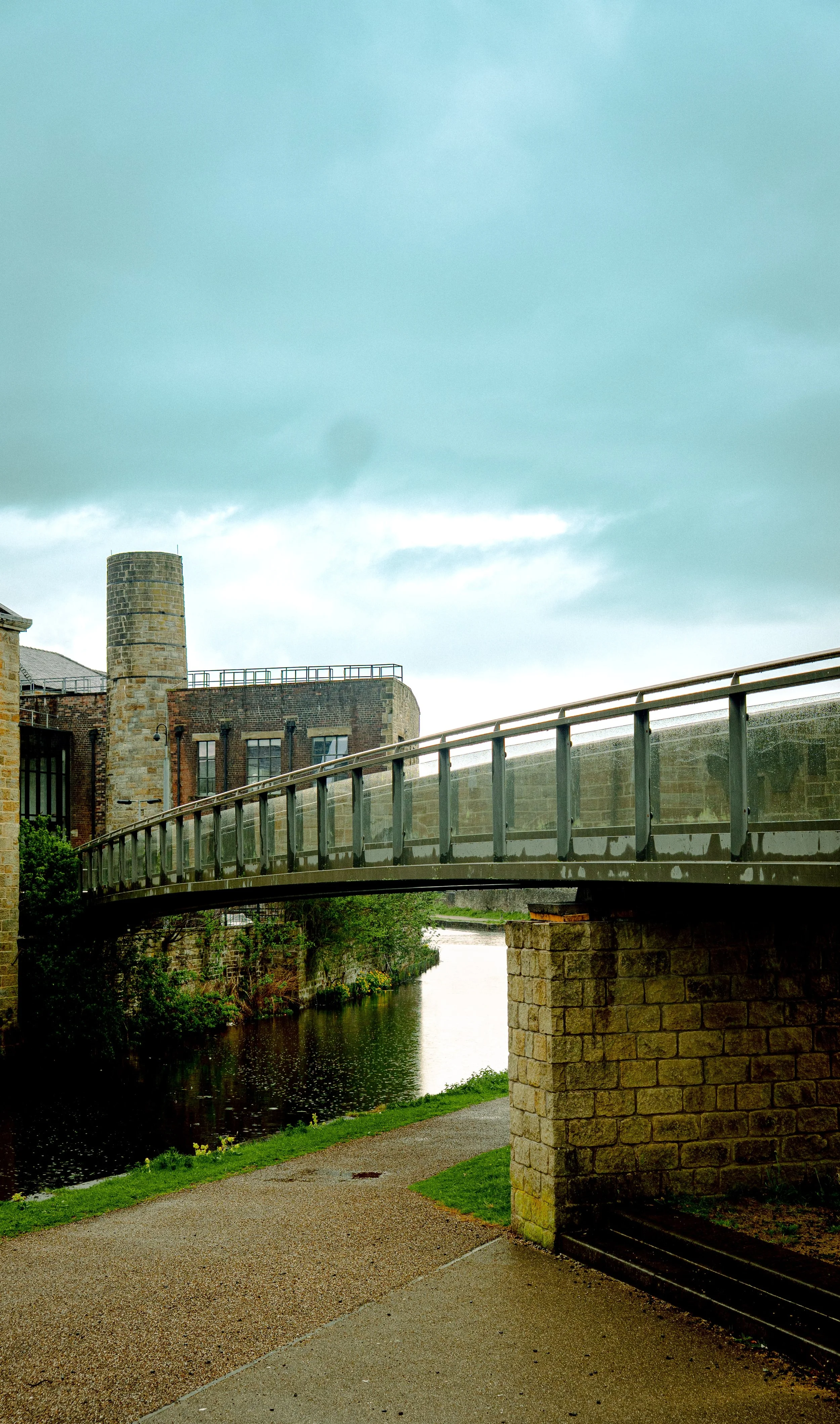 A bridge over a small river with an industrial-style building and chimney in the background, under a cloudy sky.