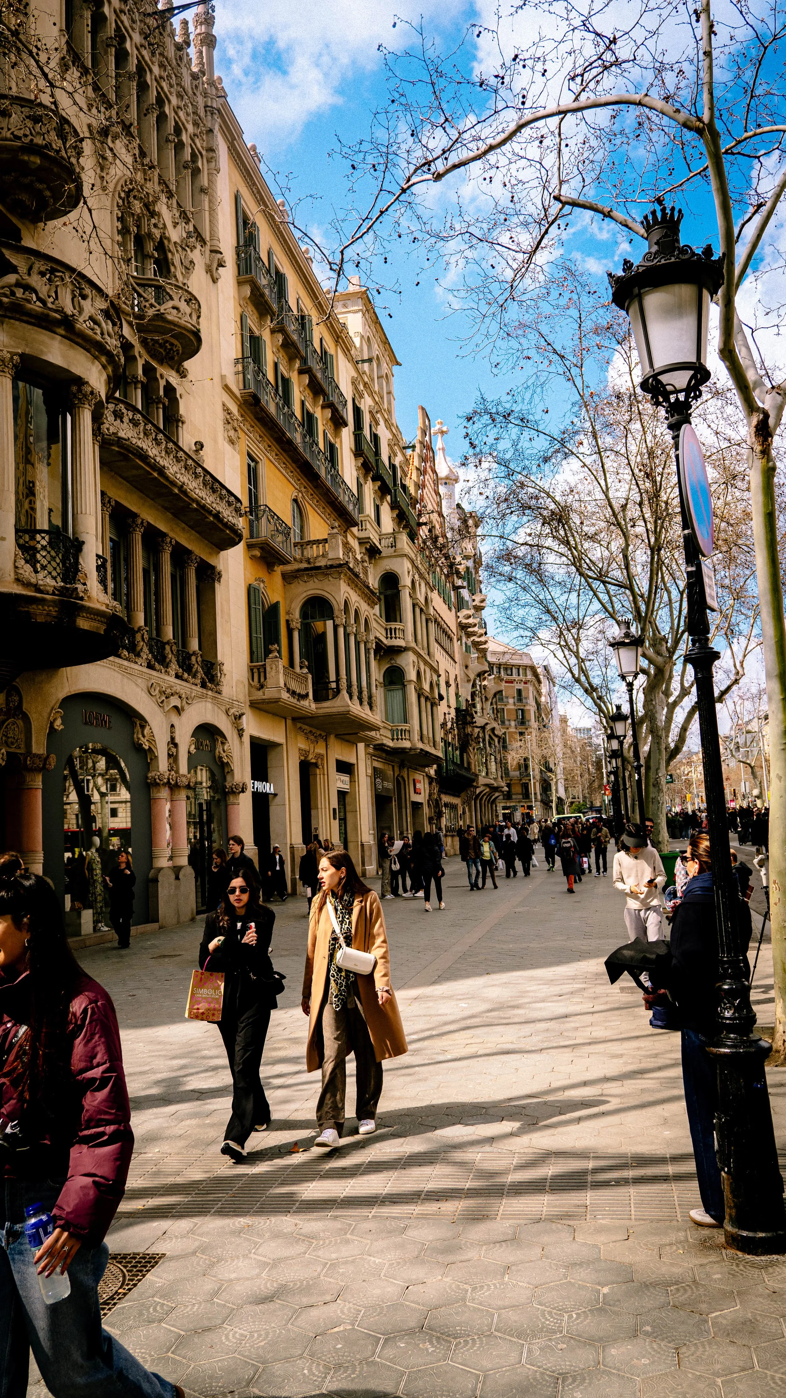A busy city street with ornate buildings, leafless trees, street lamps, and pedestrians walking on the sidewalk.