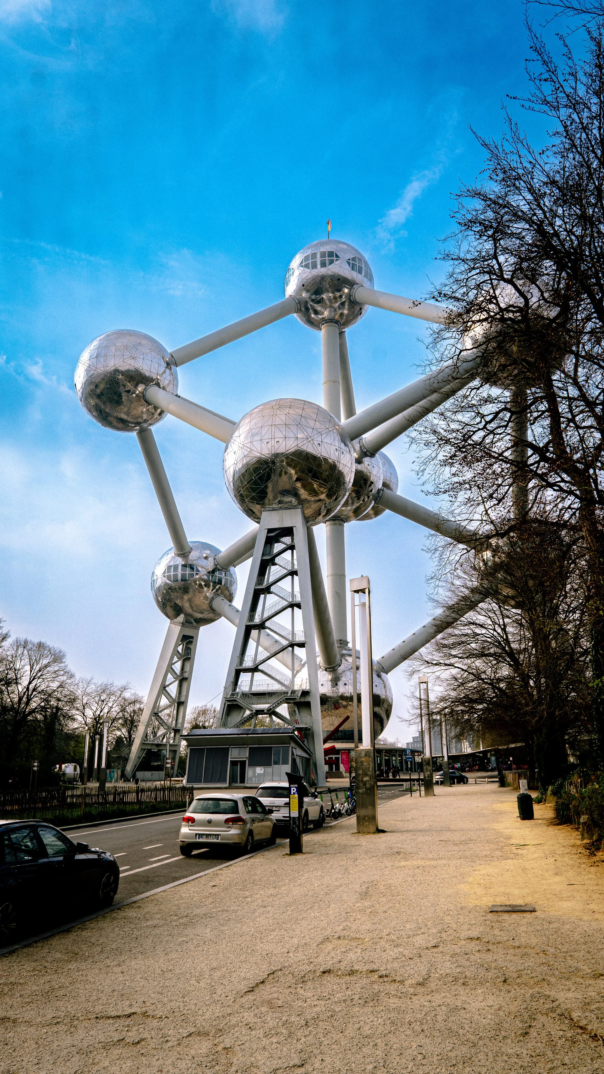 The Atomium, a large metallic structure resembling a model of an atom with interconnected spheres, located outdoors in a park setting with trees and parked cars beneath a blue sky.