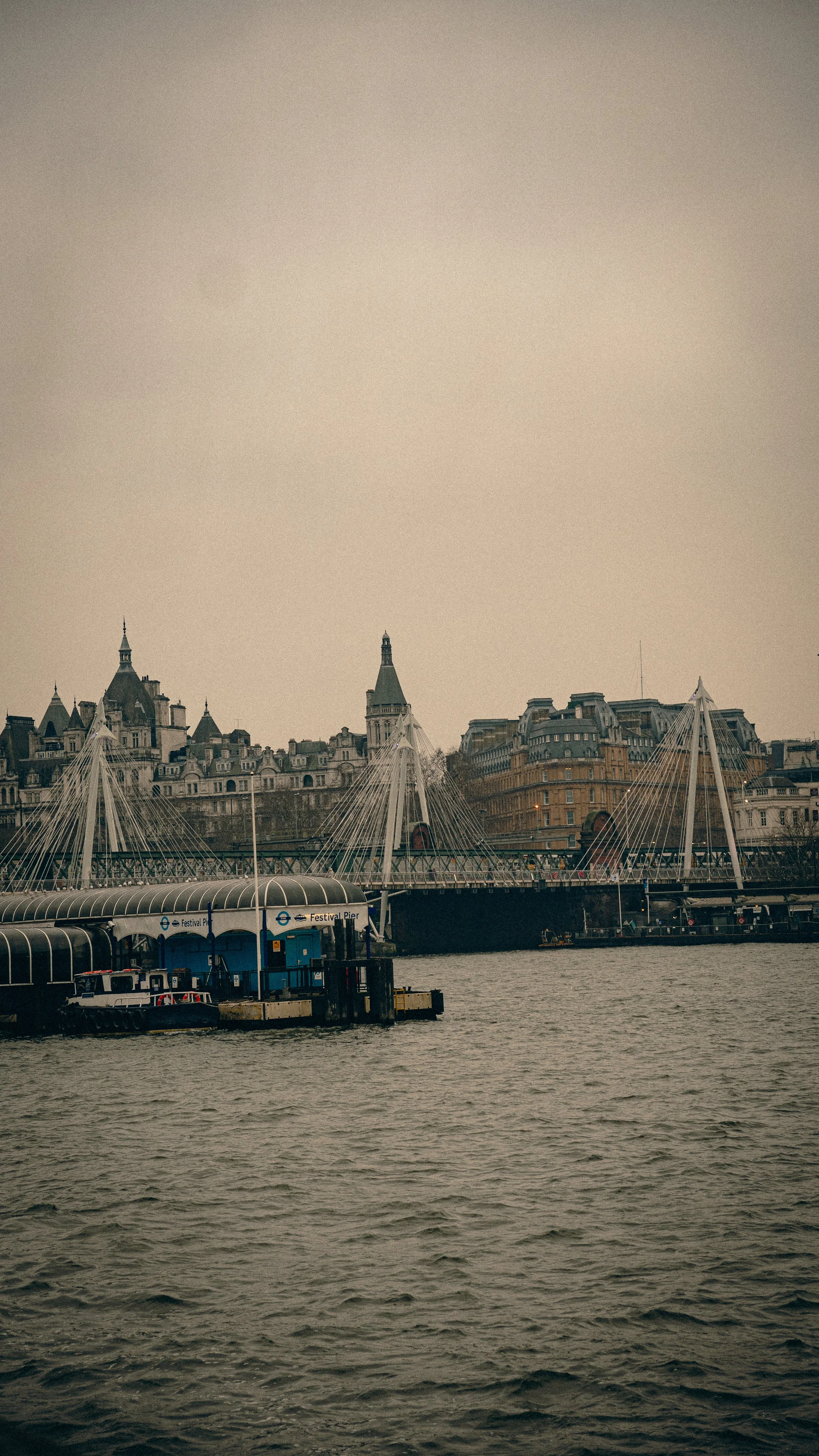 View of the riverside with a bridge and waterfront buildings in London, including the Festival Pier sign and the London Eye's structures in the background.