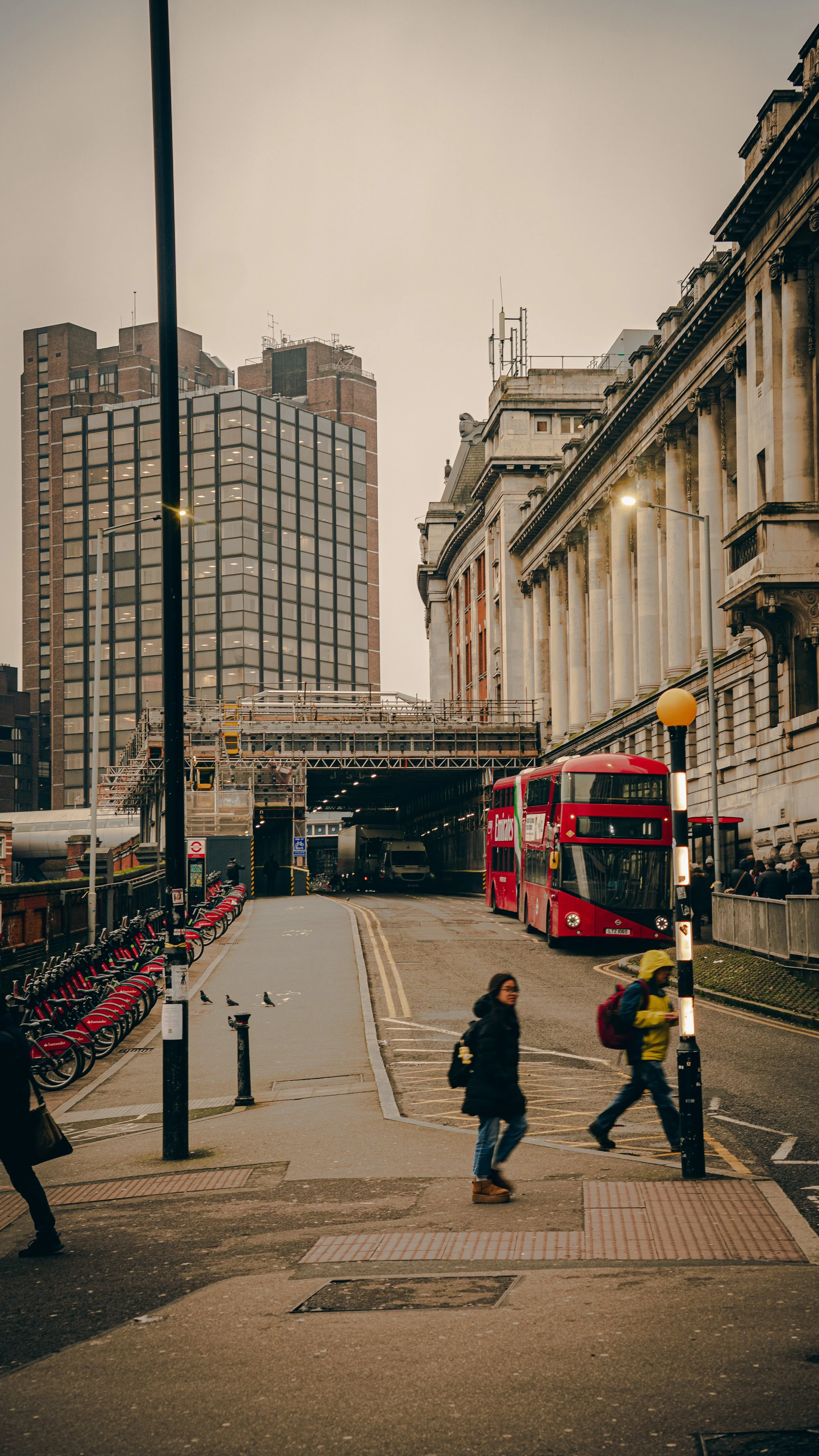 A city street scene with pedestrians crossing, a red double-decker bus, and tall buildings in the background.