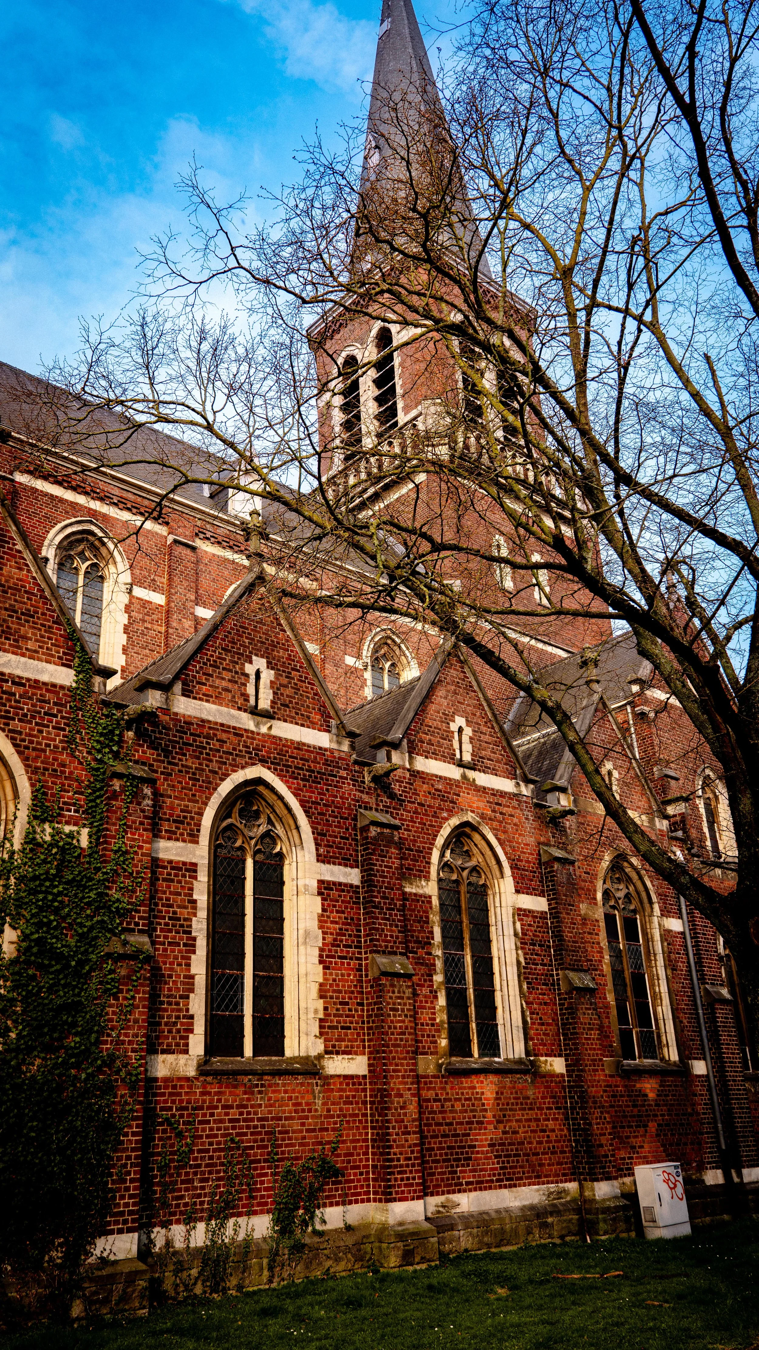 A brick church with pointed arched windows and a tall steeple, seen through leafless tree branches against a blue sky.