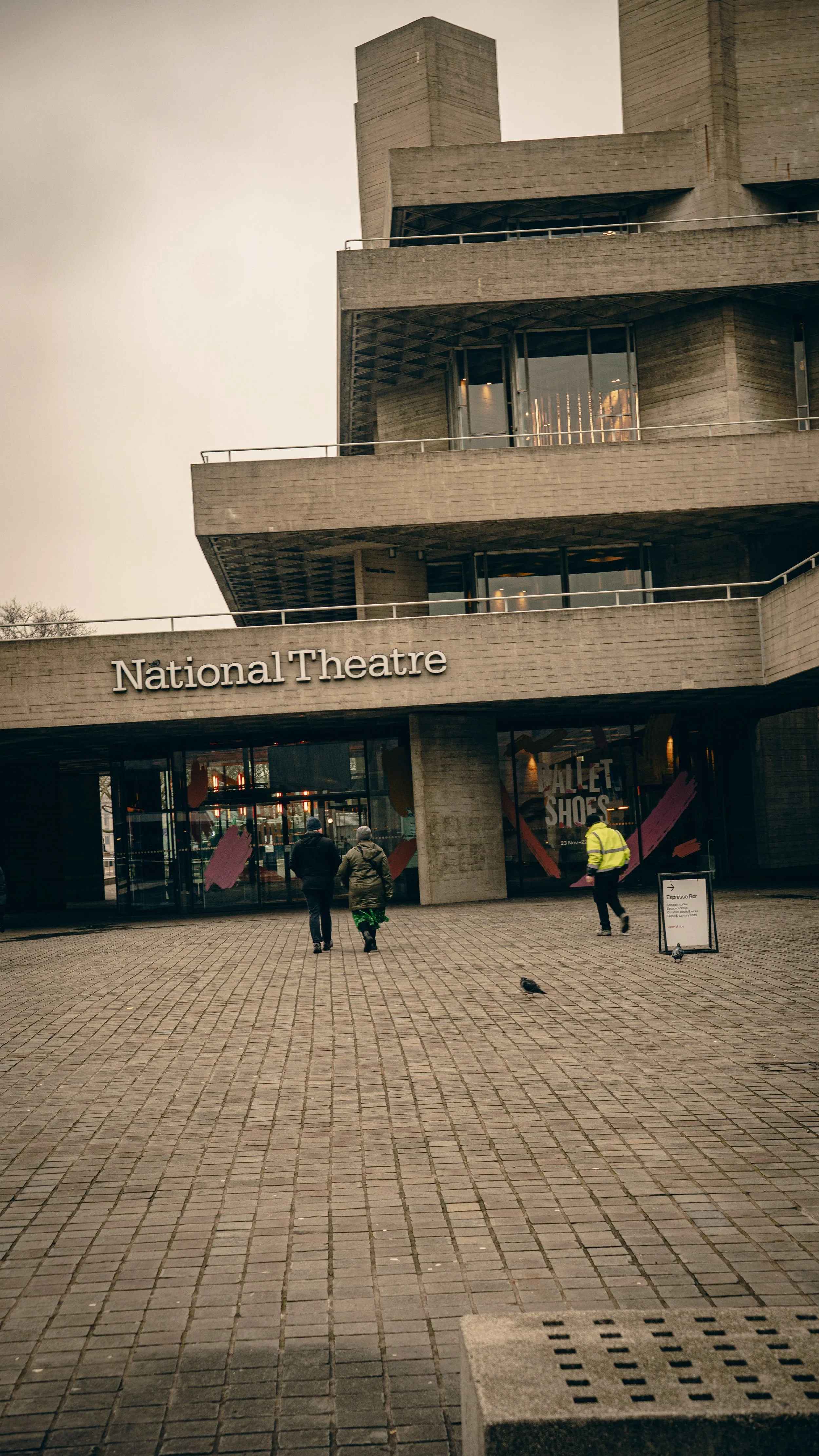 Exterior view of the National Theatre building with three people walking towards the entrance, pigeons on the pavement, and a signboard on the sidewalk.