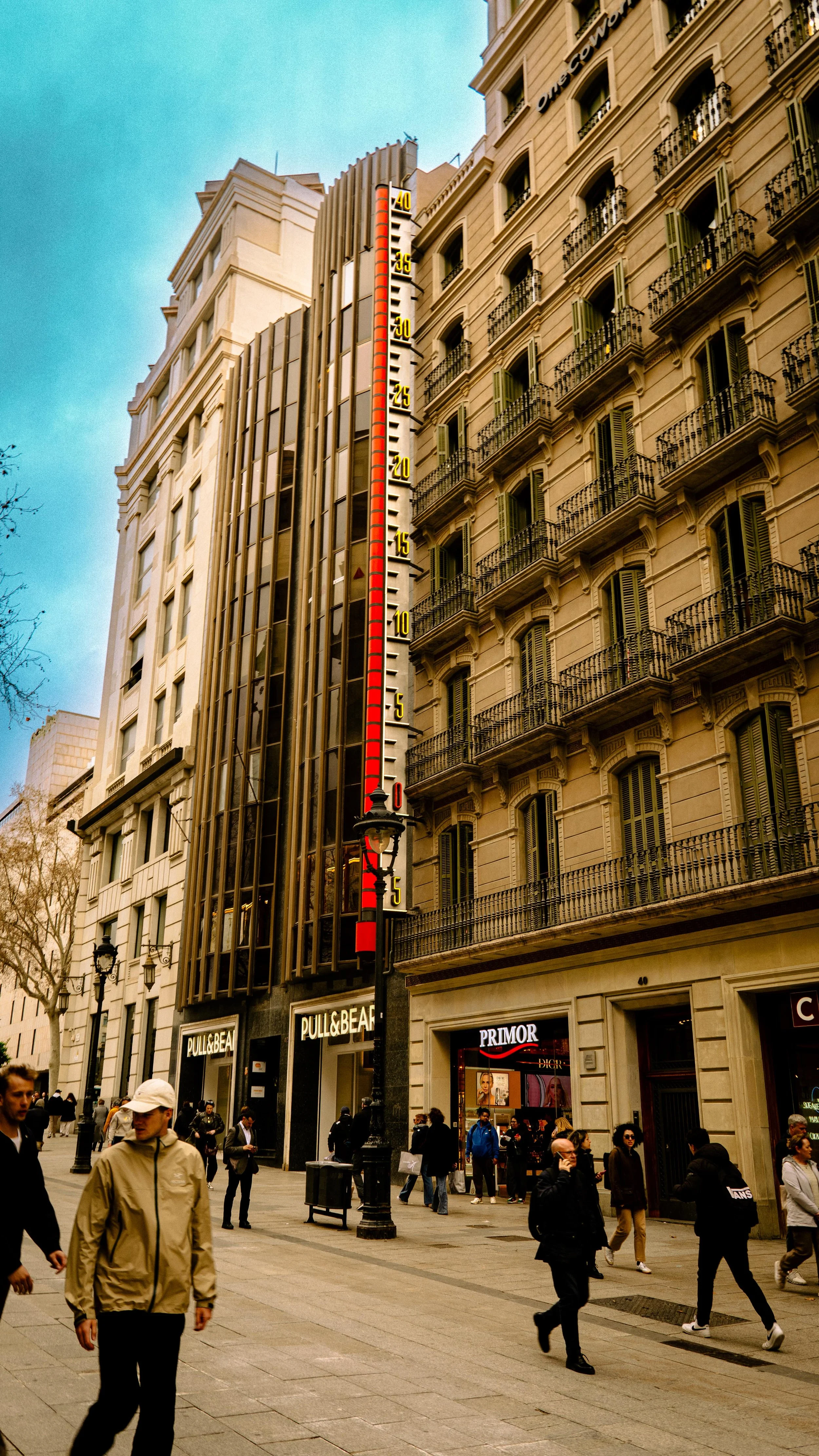 City street scene with pedestrians in front of buildings, including a tall structure with a large red and white thermometer on its facade and storefronts like Pull & Bear and Primor.