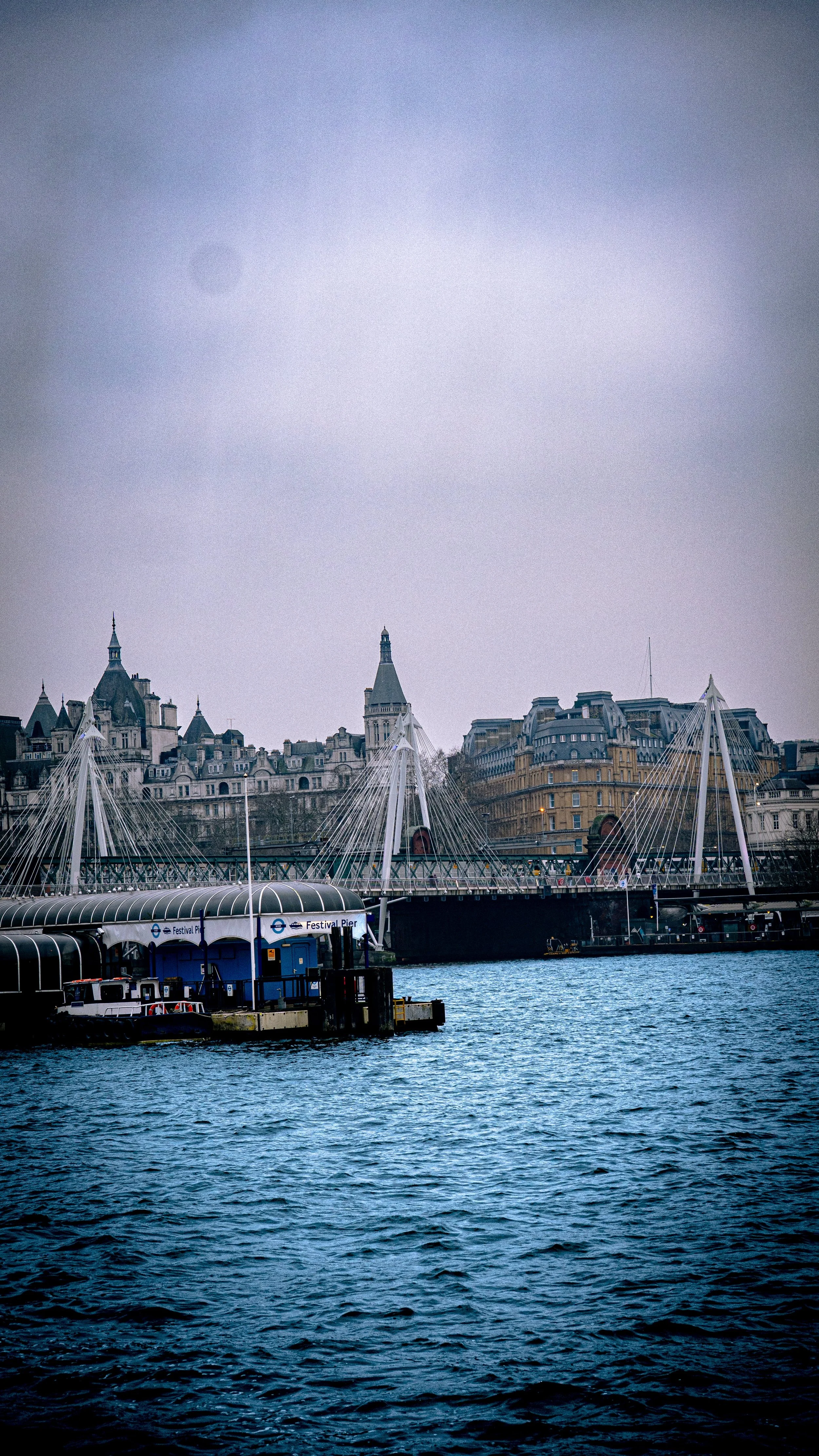 View of the Thames River with the Festival Pier on the riverbank and the Millennium Bridge in London, toward historic buildings in the backdrop under cloudy sky.