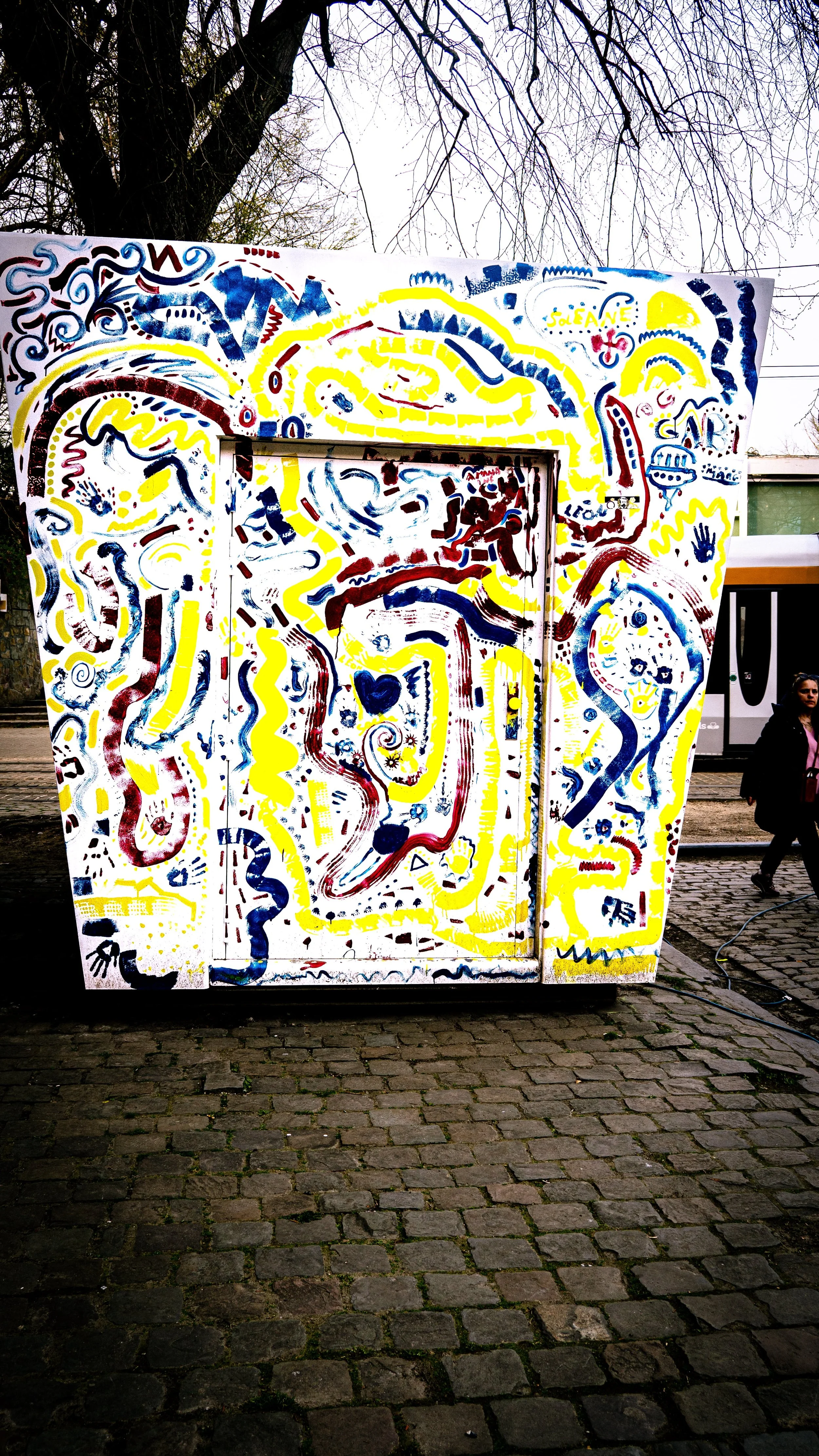 Colorful abstract mural with swirling lines and shapes in yellow, red, and blue on a white background, painted on an outdoor structure with cobblestone ground.
