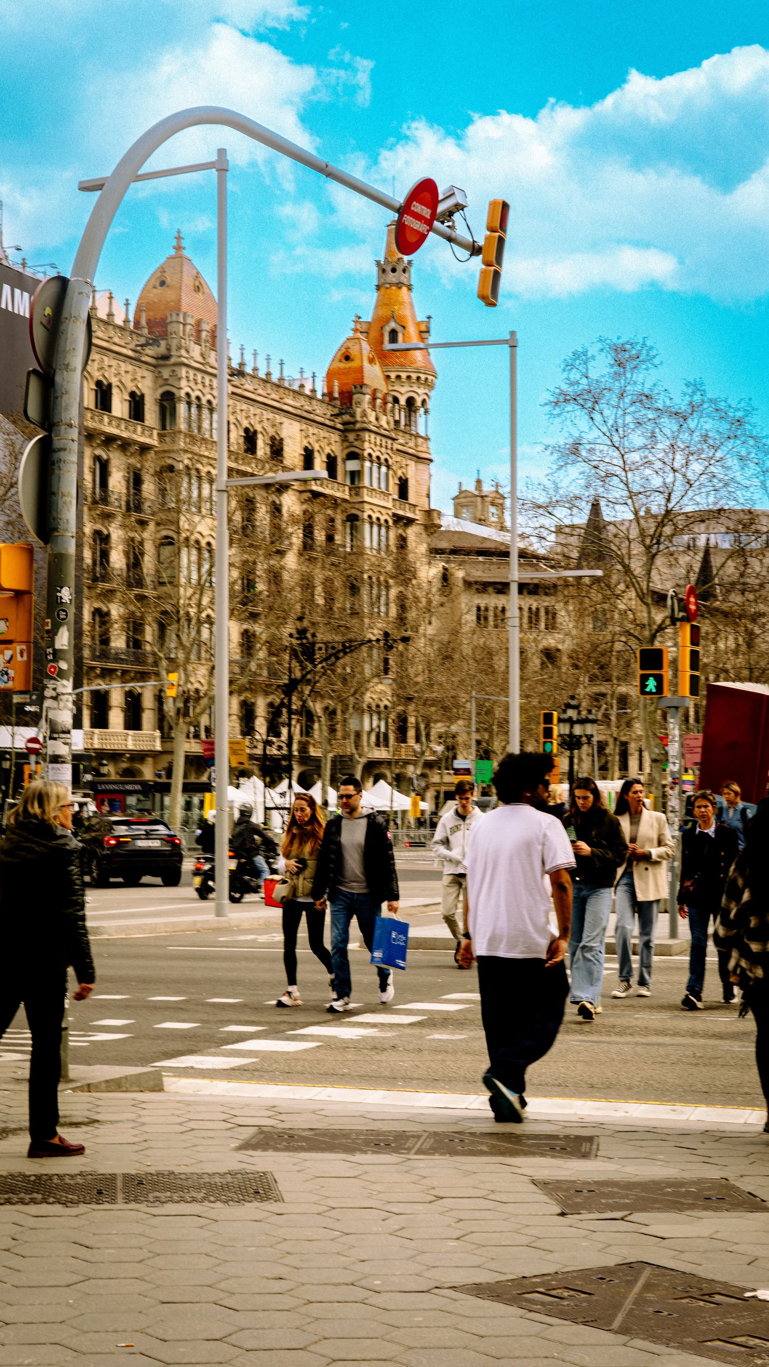 A busy city street crosswalk with pedestrians walking across. In the background, there is a historic building with architectural details and turrets, and traffic lights overhead. The sky is partly cloudy and there are leafless trees along the street.