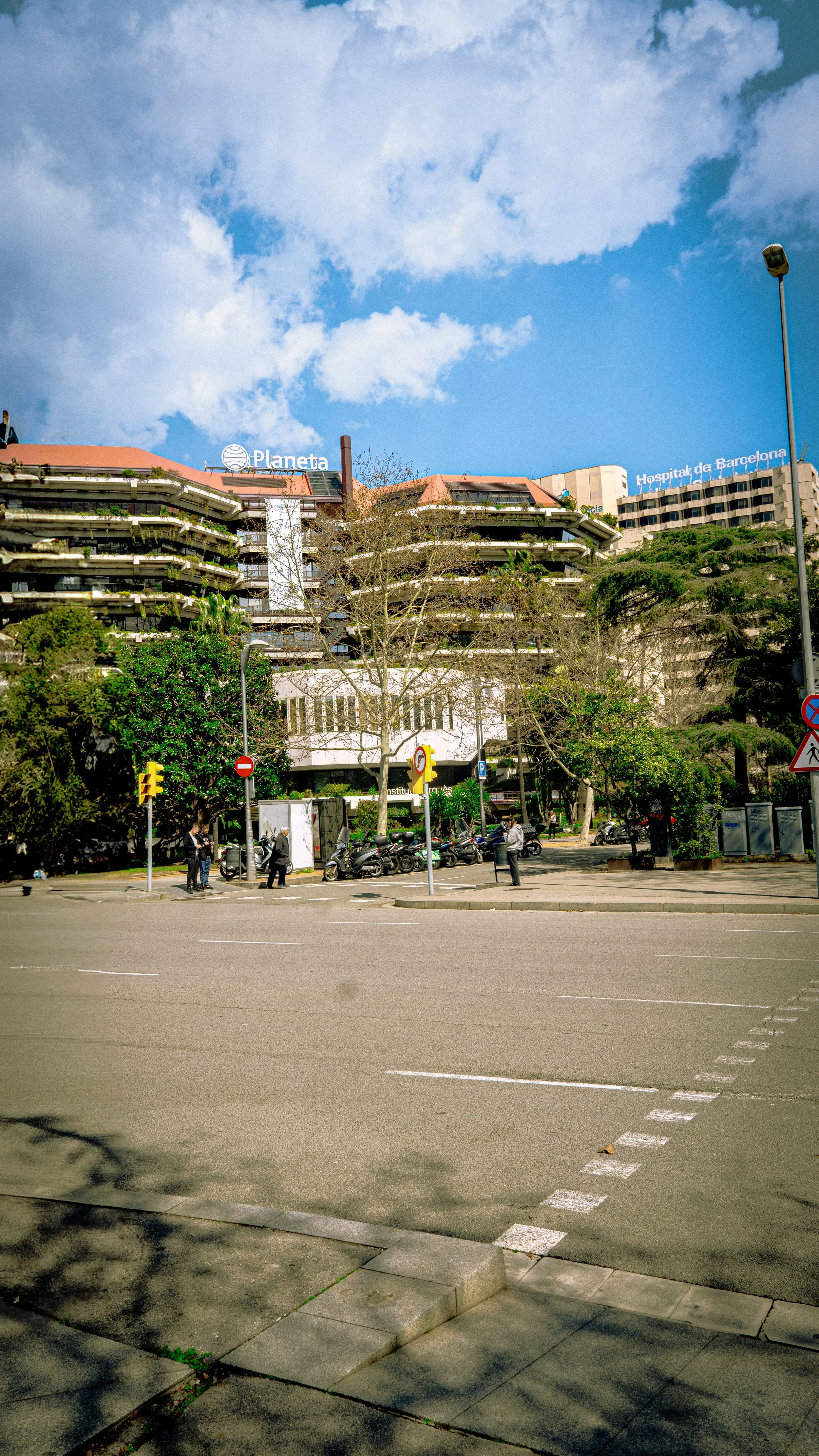 Street view in Barcelona with trees, scooters, pedestrians, and buildings including Hospital de Barcelona and Planeta signs under a partly cloudy sky.