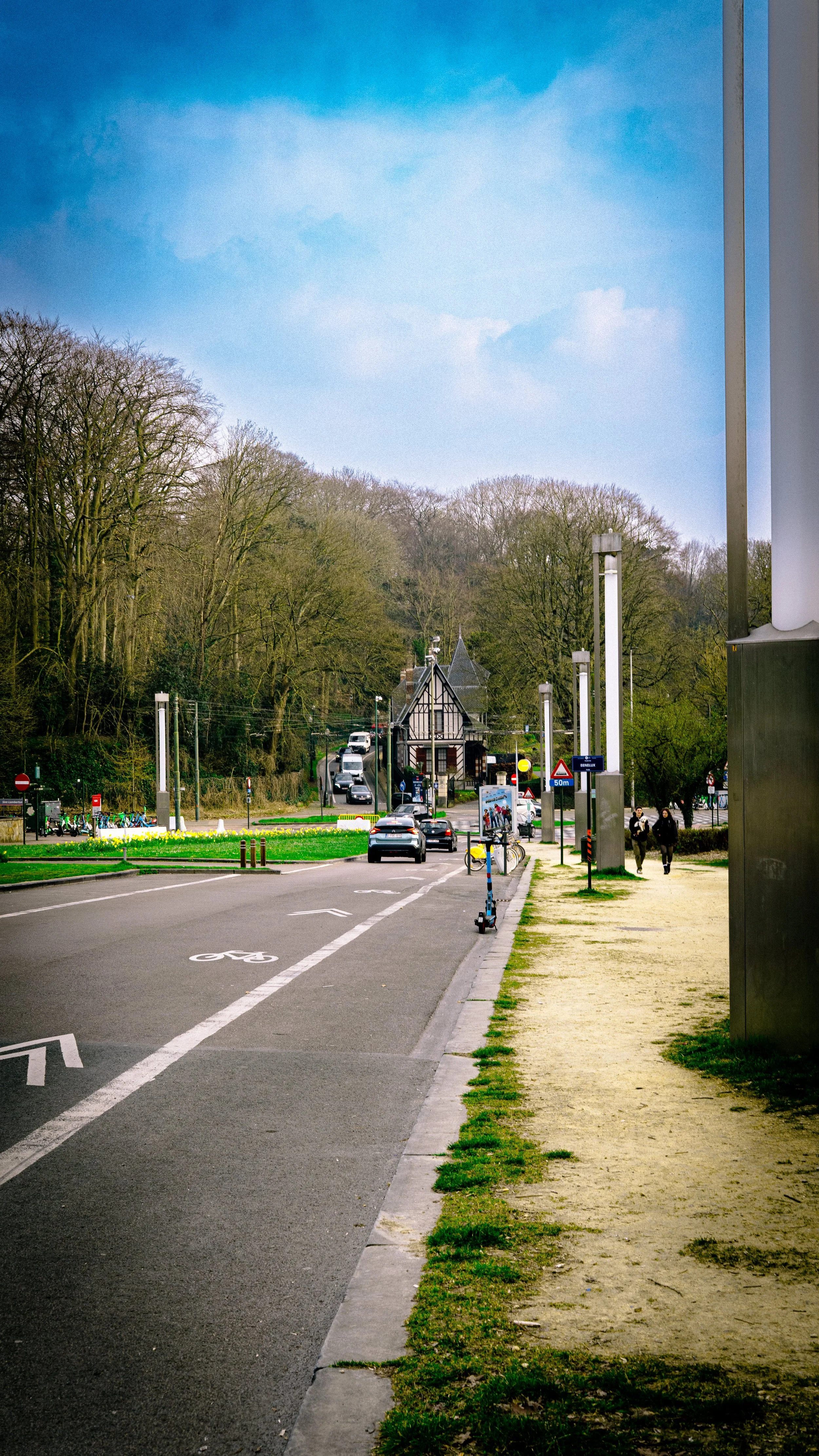 A scenic street scene with a bicycle lane and sidewalk, some pedestrians, trees, parked bicycles, and a small building in the distance under a partly cloudy sky.