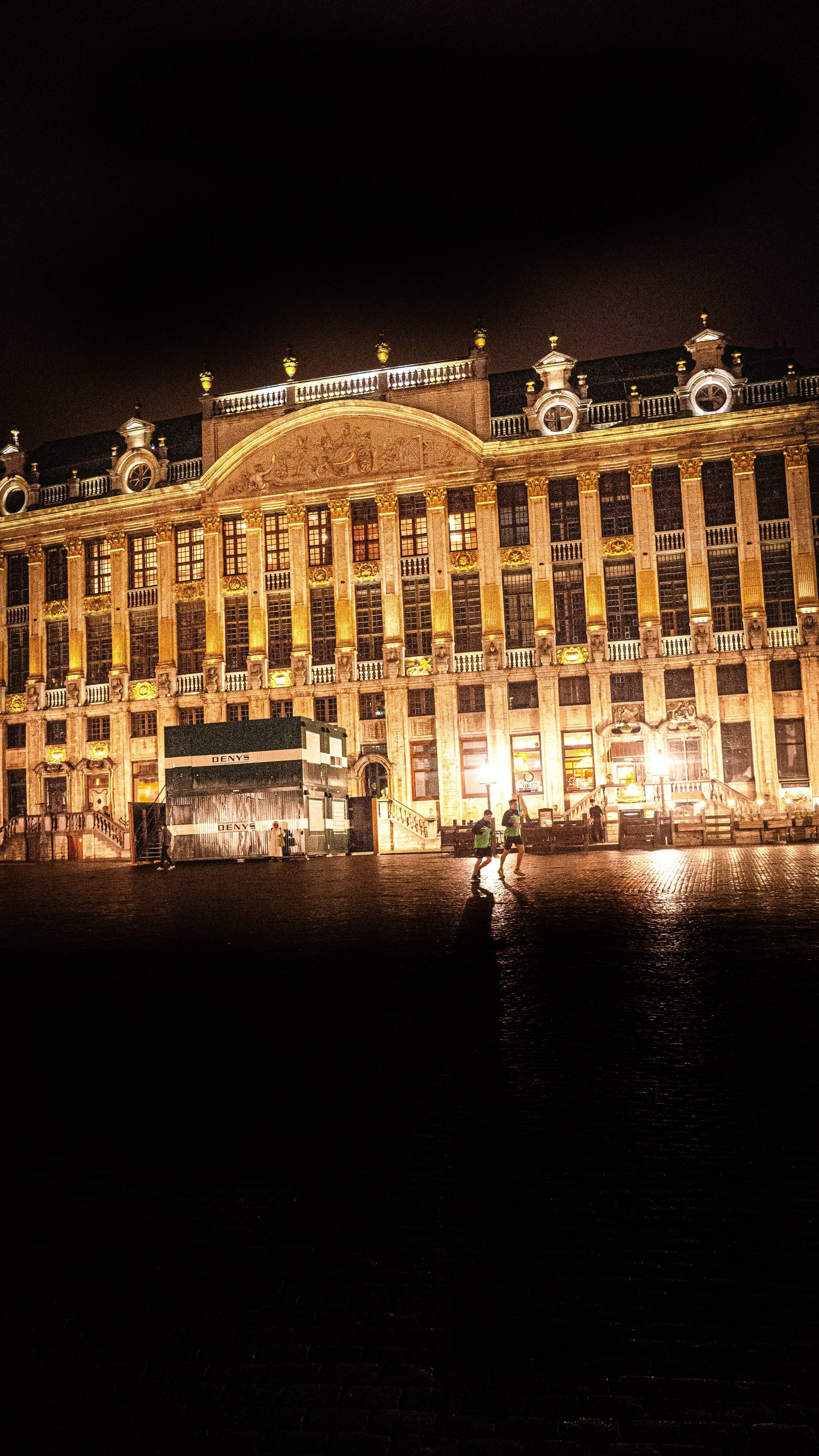 A large, ornate building illuminated at night, with a few people running in the wet cobblestone street in front.