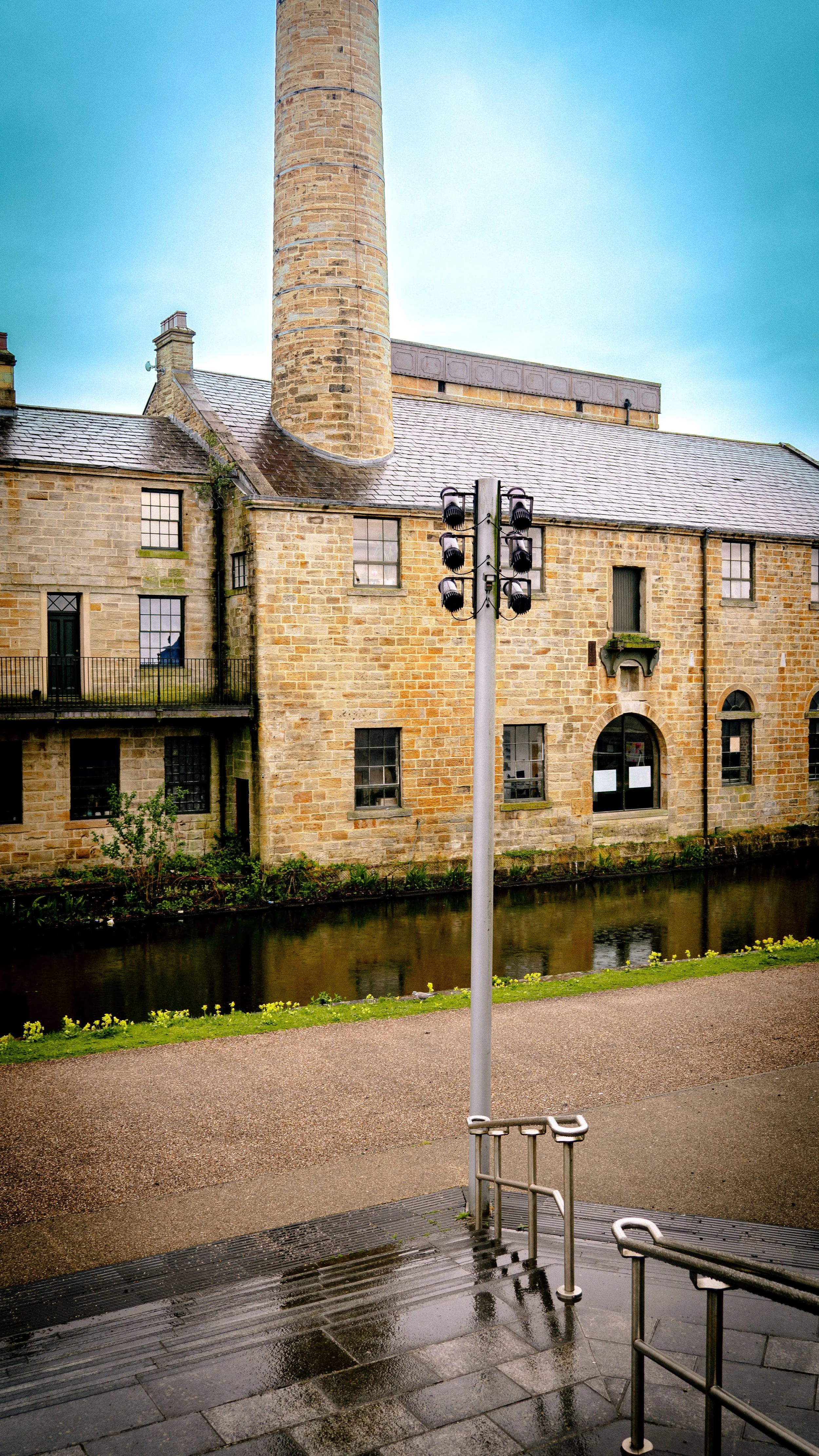 Historic stone building with a rounded chimney, windows, a balcony, a small waterway, a lamp post, and rain on the pavement.