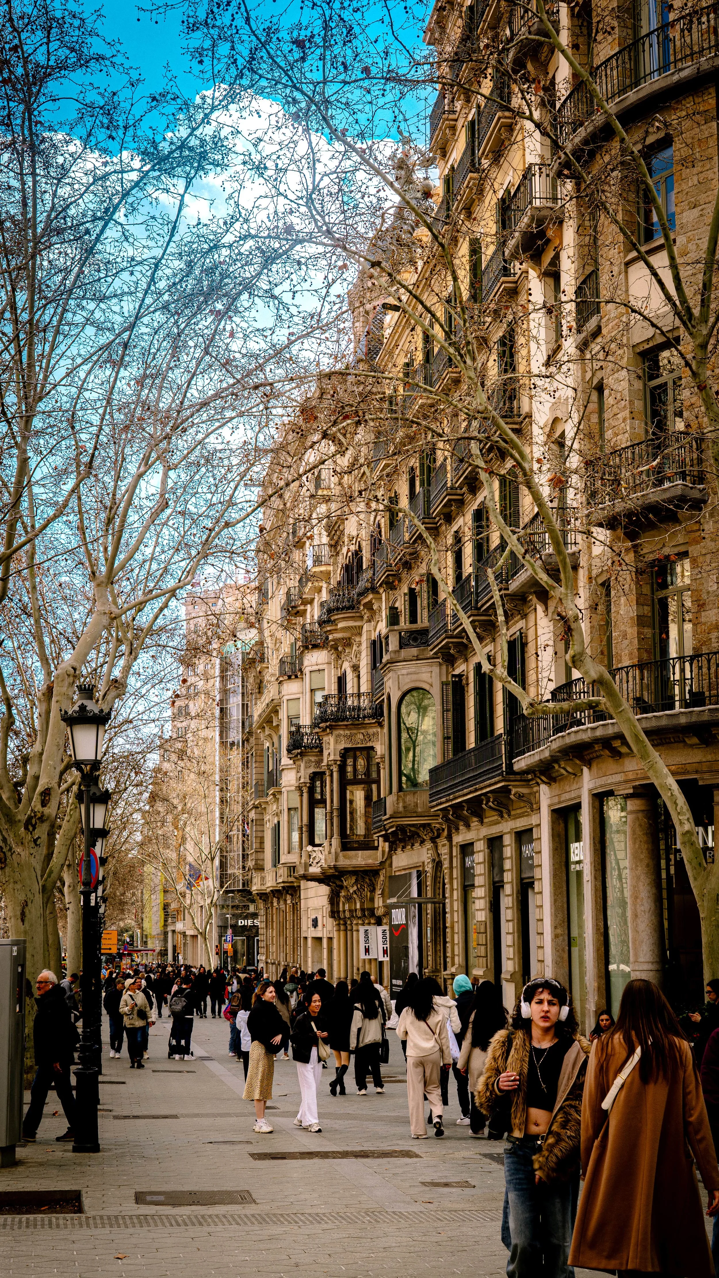 Crowded city street with people walking and shopping, surrounded by historic buildings with balconies and leafless trees, under a blue sky.