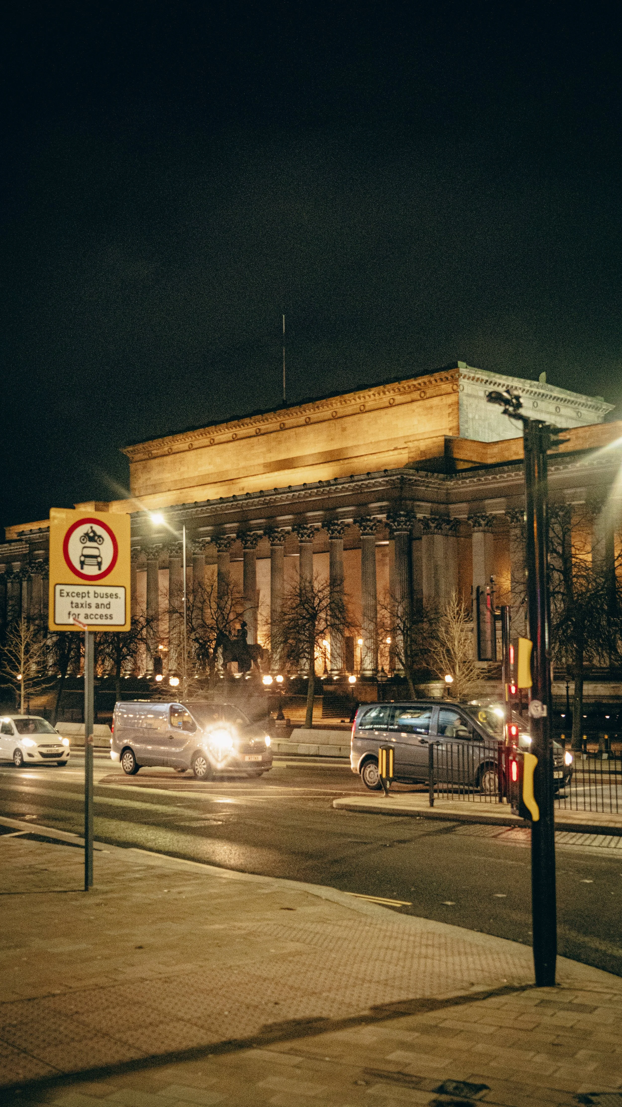 Nighttime view of a historic building with classical columns, illuminated by exterior lights. In the foreground, a busy street with cars and a traffic light. A sign indicates parking restrictions except for buses, taxis, and for access.