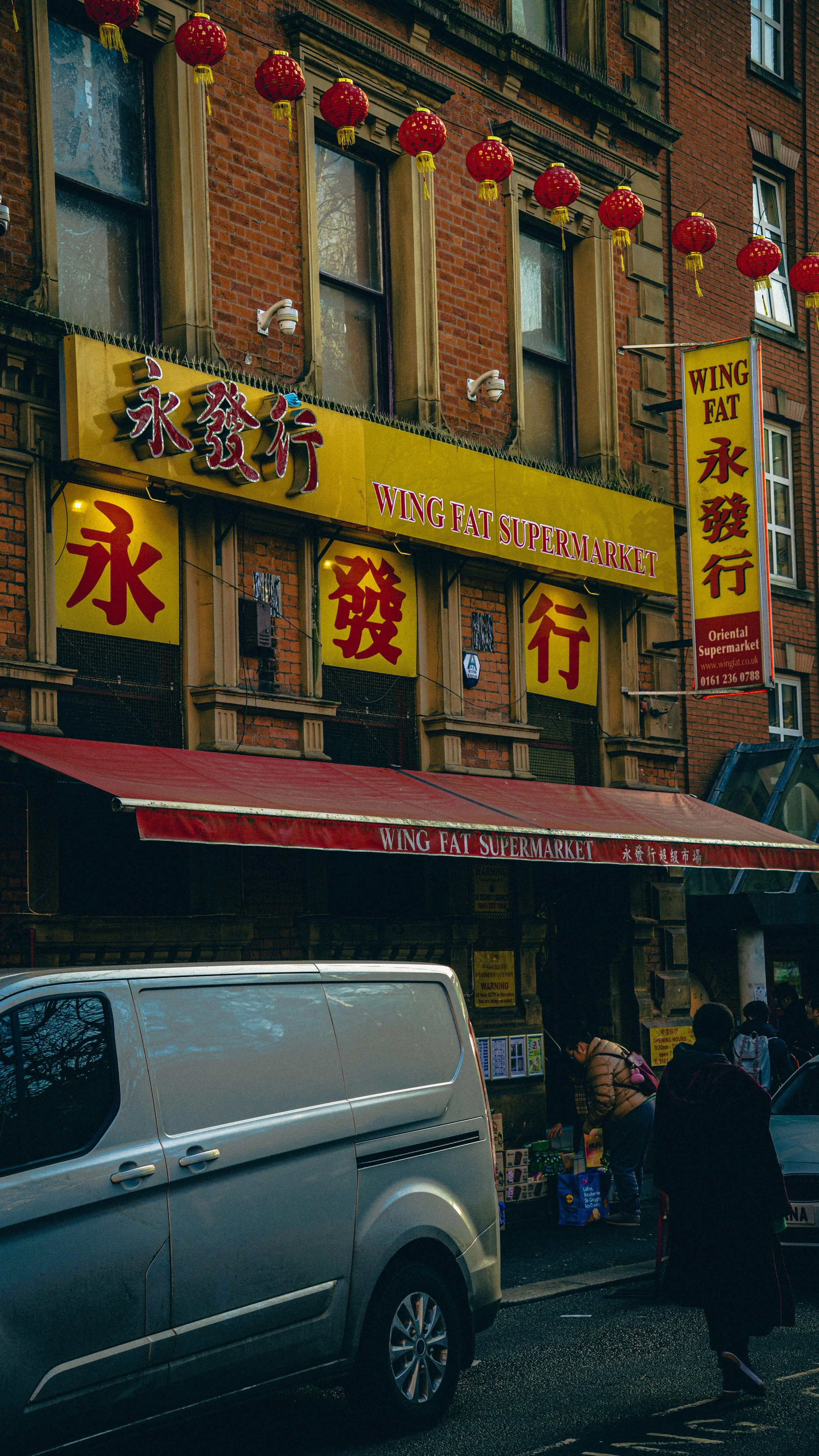 Street scene outside Wing Fat Supermarket with red lanterns hanging overhead, yellow signage with Chinese characters, people shopping and walking past parked cars.