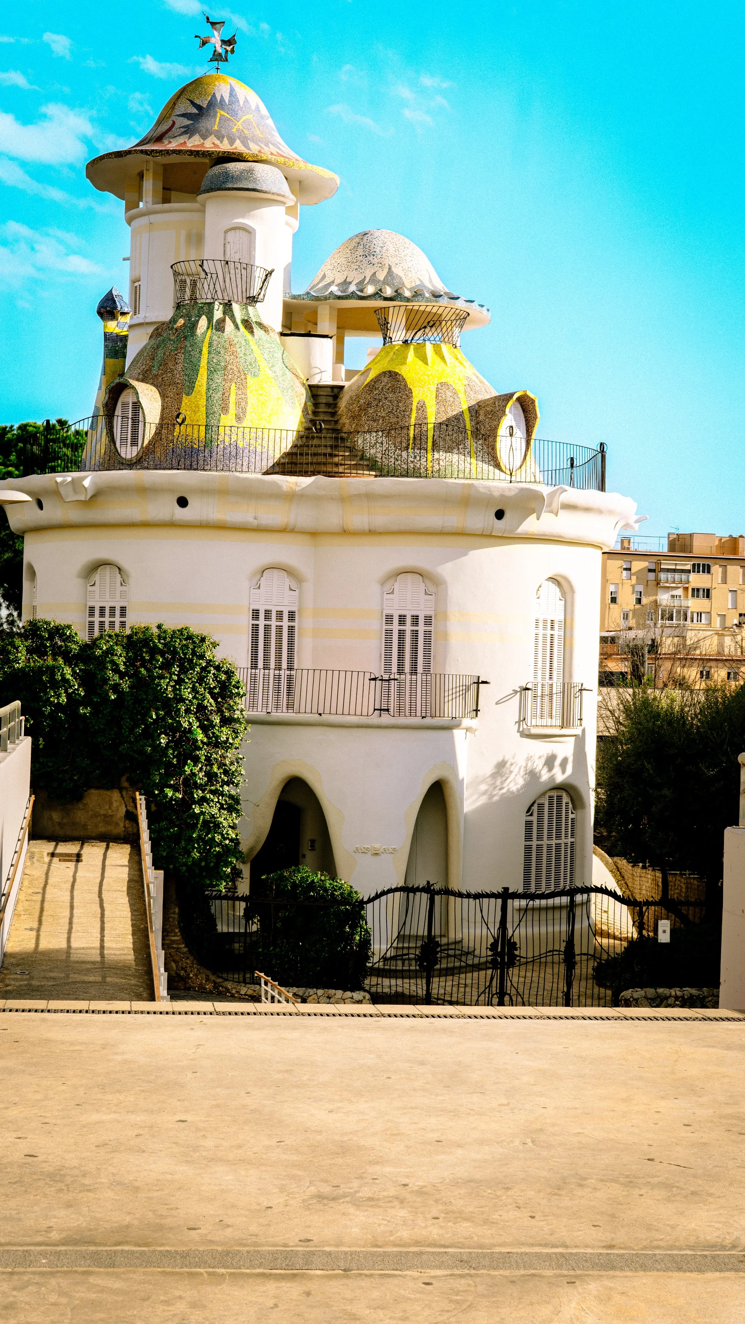 Colorful whimsical house with mosaic roof, rounded windows with shutters, and decorative stairs, set against a clear blue sky.