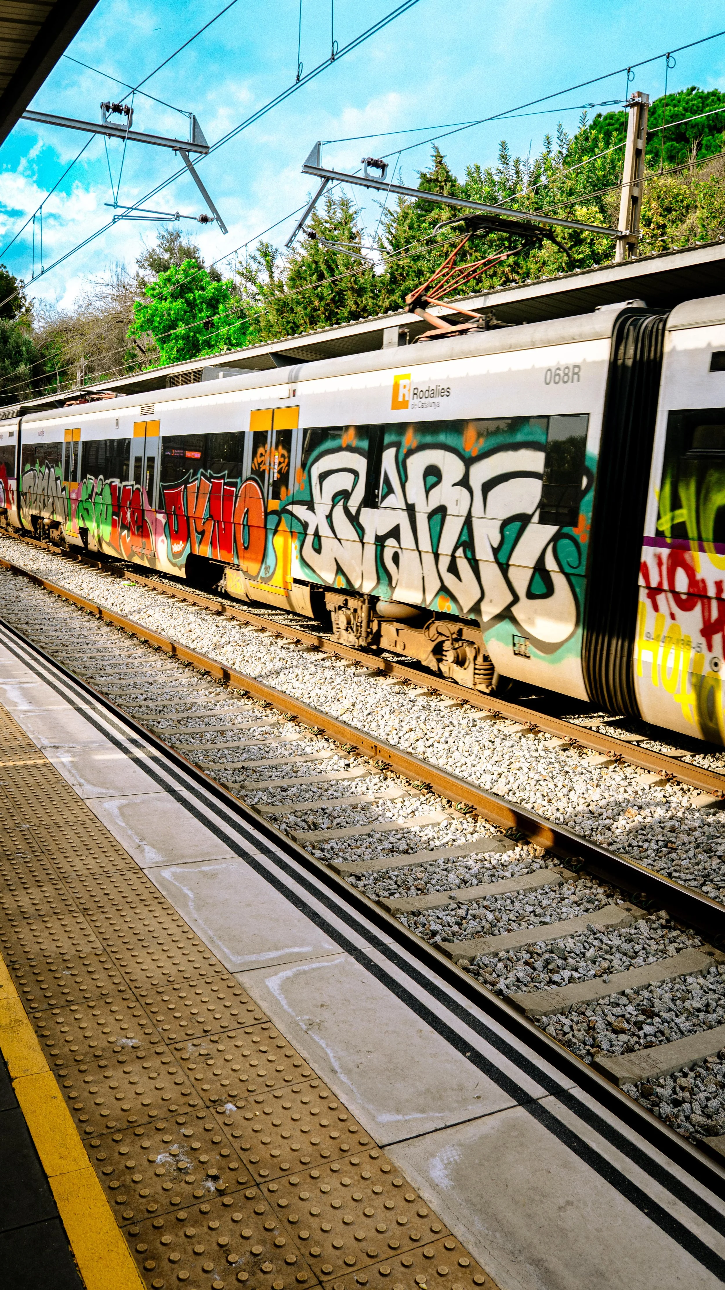 A train with graffiti on its side at a train station platform, with trees and a blue sky in the background.