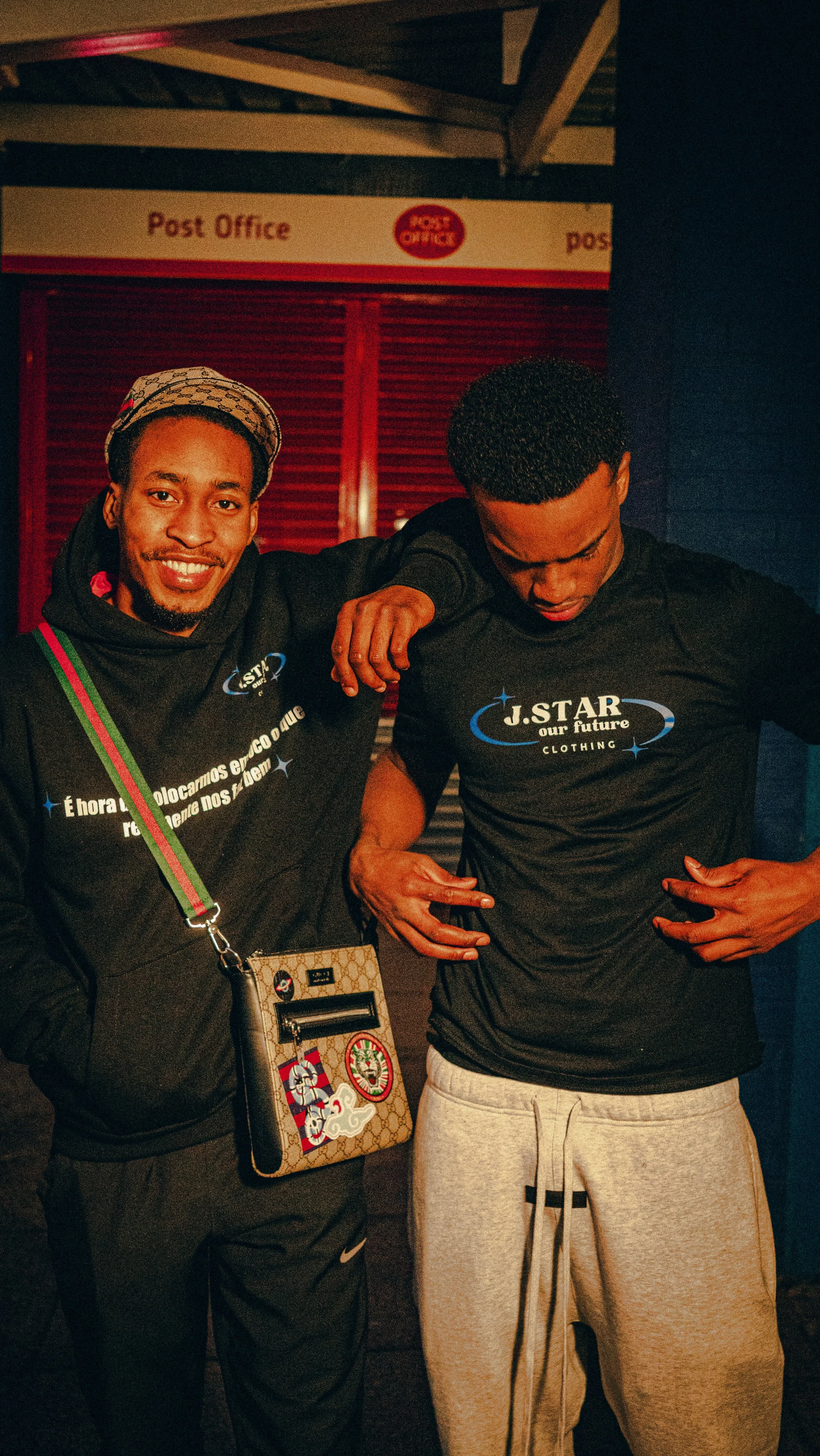 Two men posing cheerfully in front of a post office sign at night. One man is smiling, wearing a hoodie with a small bag, and the other is looking down, adjusting his shirt. Both are wearing black shirts with white and blue text that reads 'J.STAR ou