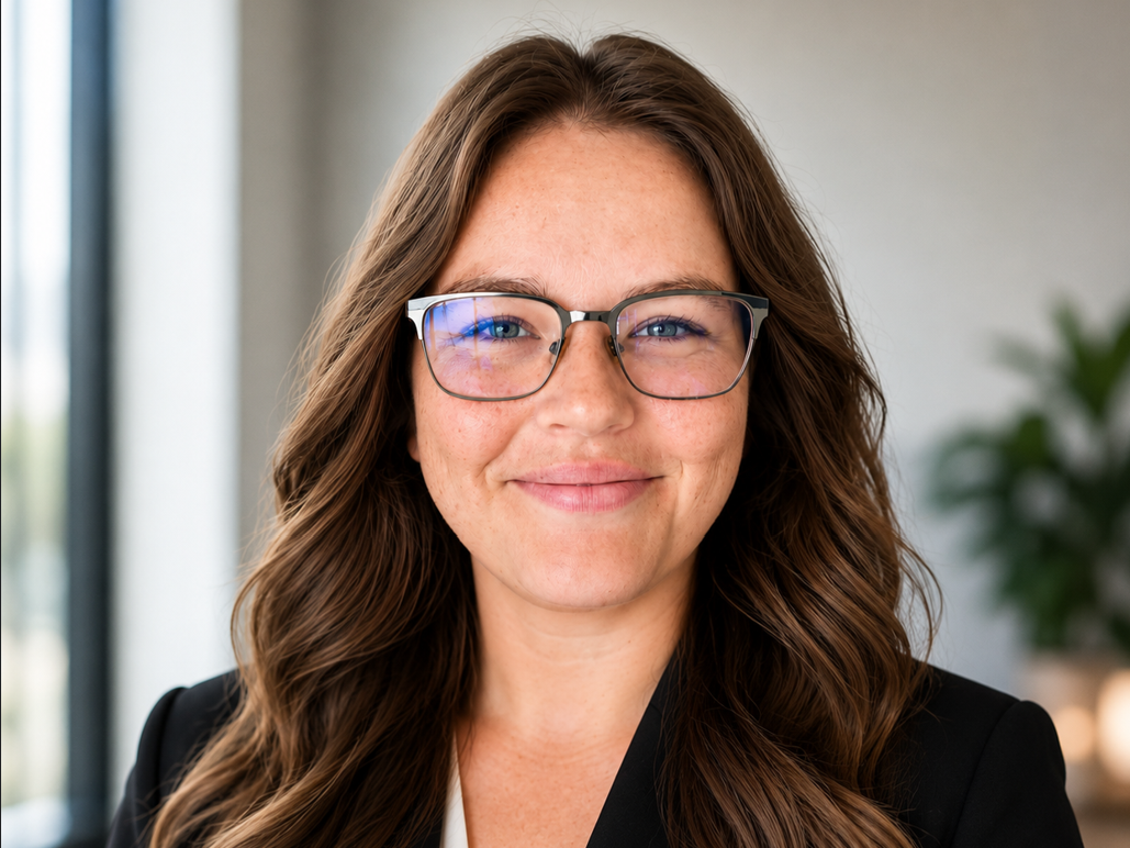 Close-up of a woman with long, wavy brown hair, wearing glasses and a black blazer, smiling confidently in an office setting.
