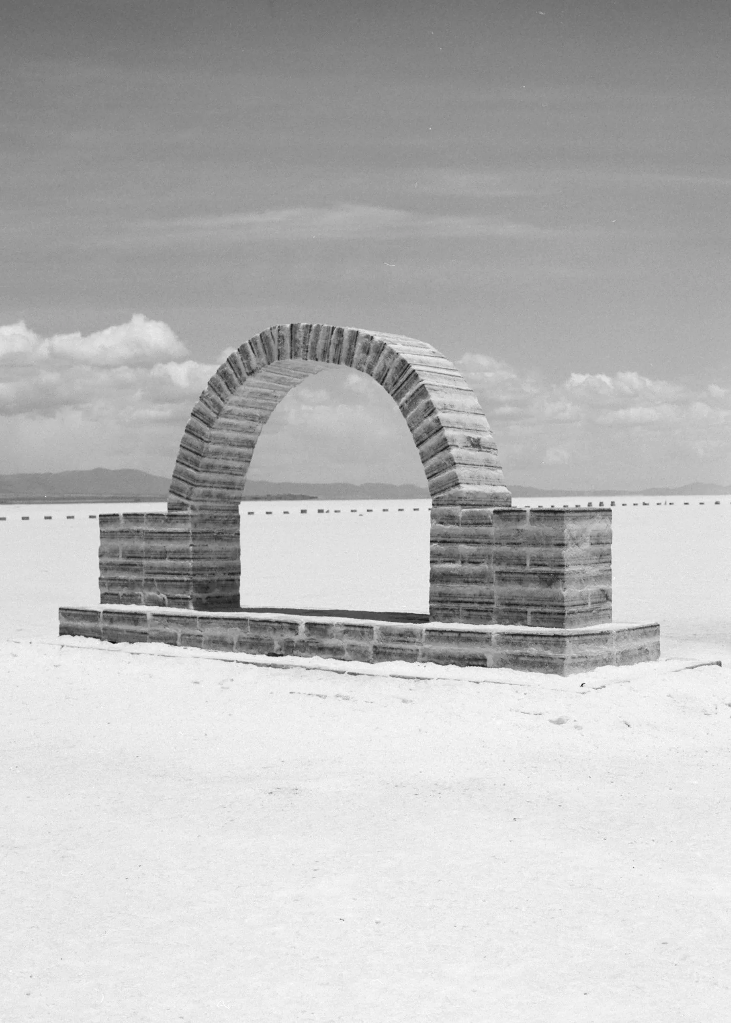A large, arched stone structure on a flat, white salt flat with a distant mountain range and blue sky with clouds in the background.