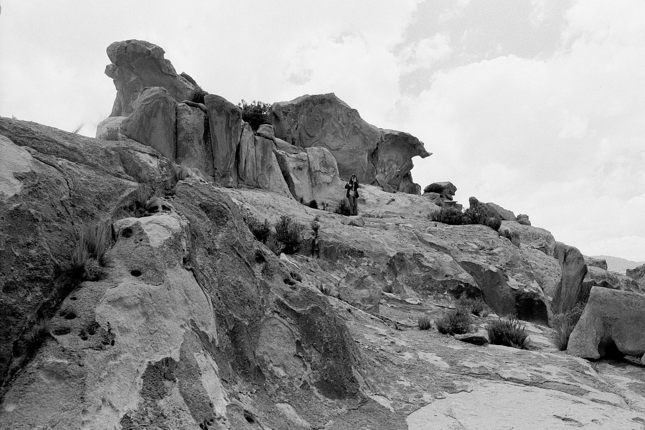 A person hiking up rocky desert terrain with large rock formations and sparse vegetation under cloudy skies.