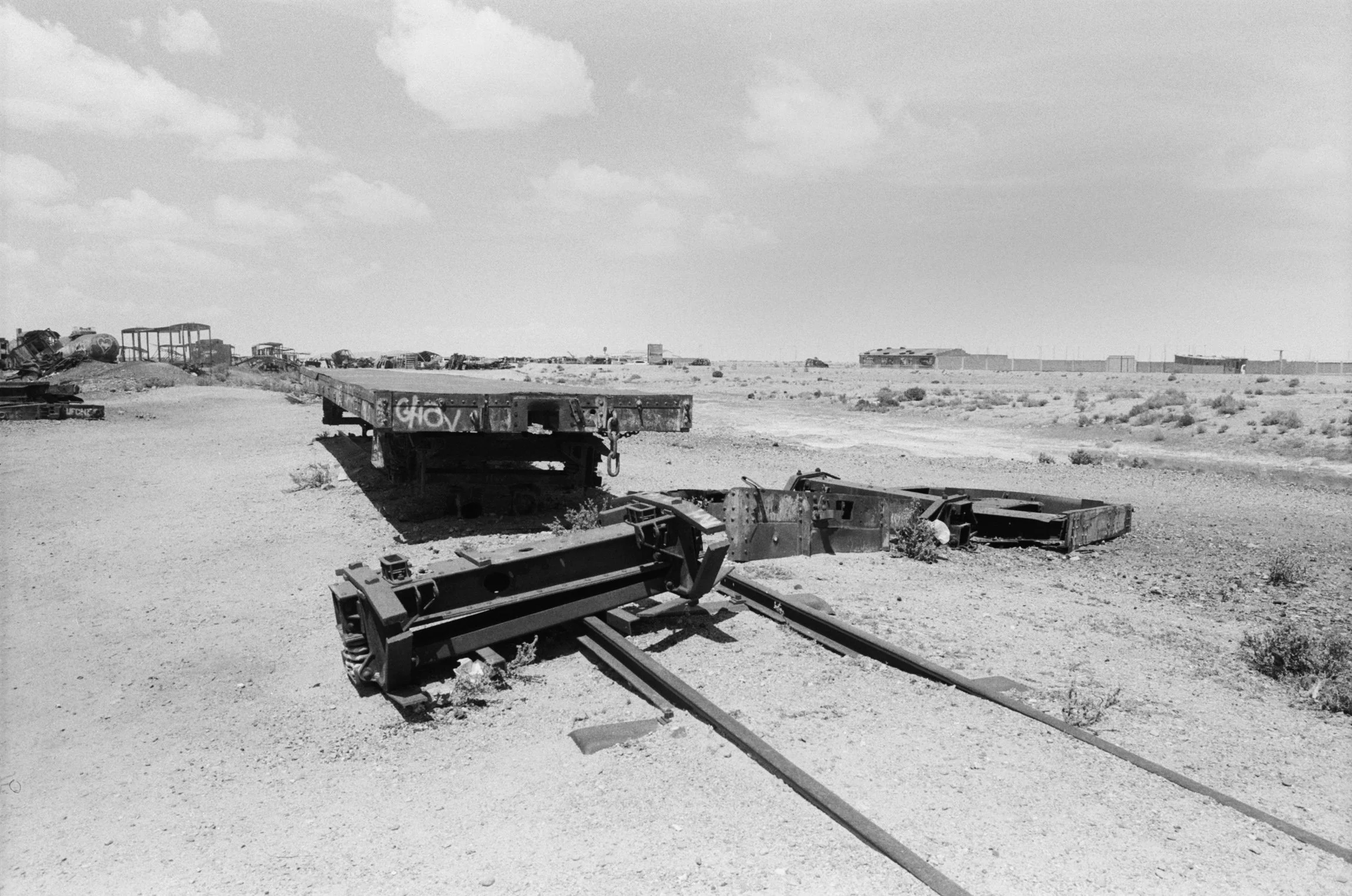Desert landscape with abandoned, rusted train track and train cars in the distance, under a partly cloudy sky.