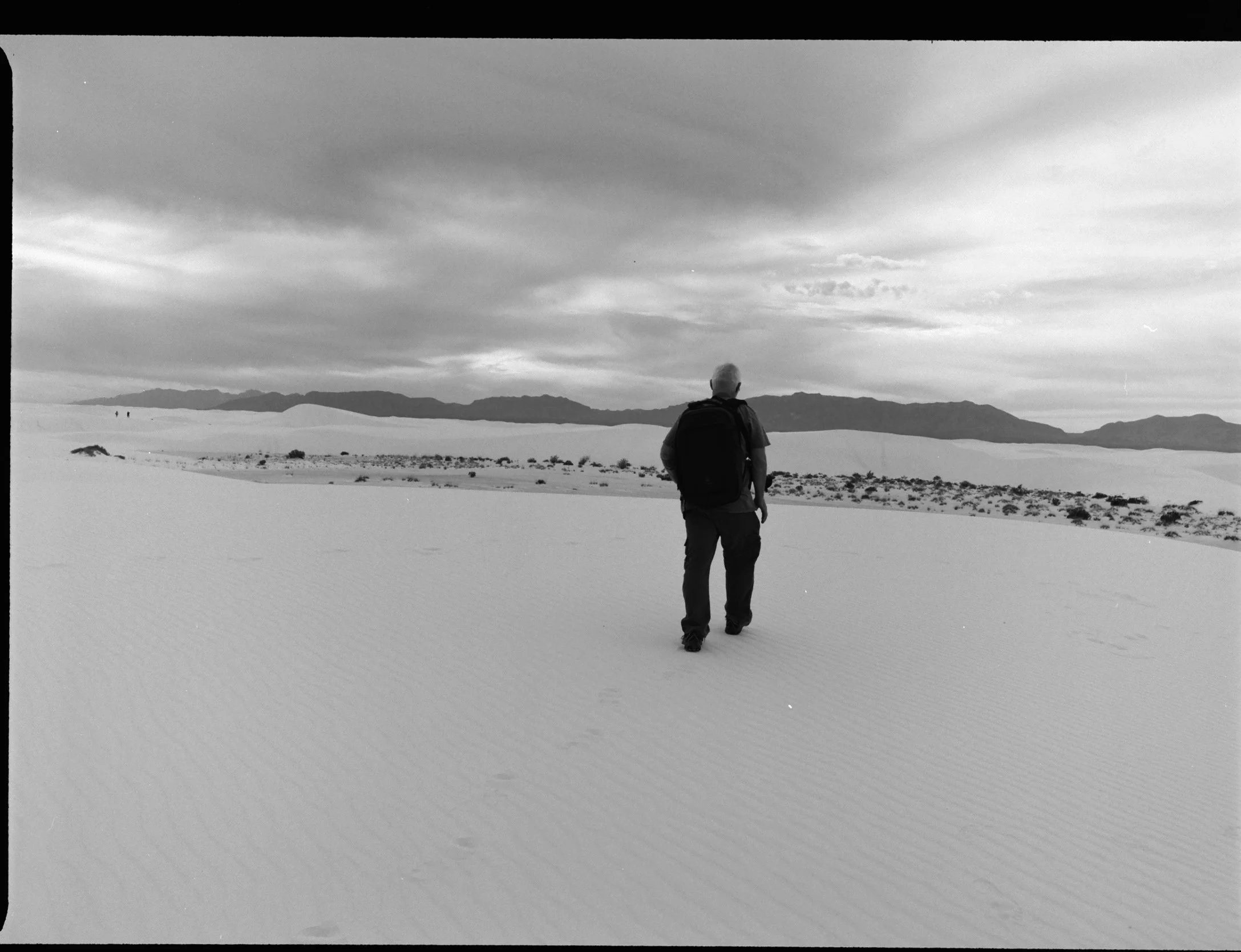 A person with gray hair, wearing a backpack, walking in a vast desert landscape with sand dunes and mountains in the background, under a cloudy sky.