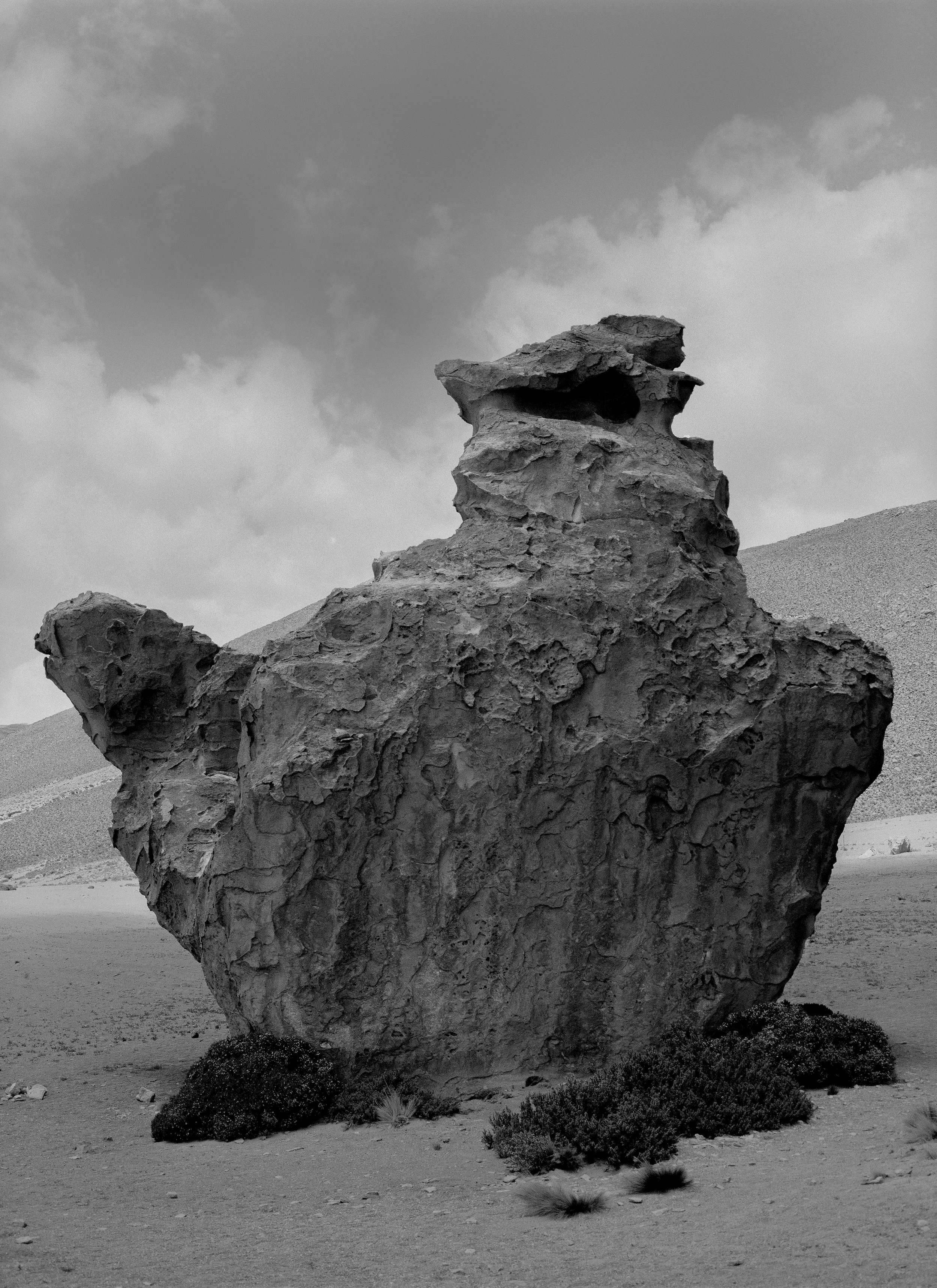 A large, weathered rock formation in a desert landscape with shrubs and mountains in the background, under a cloudy sky.