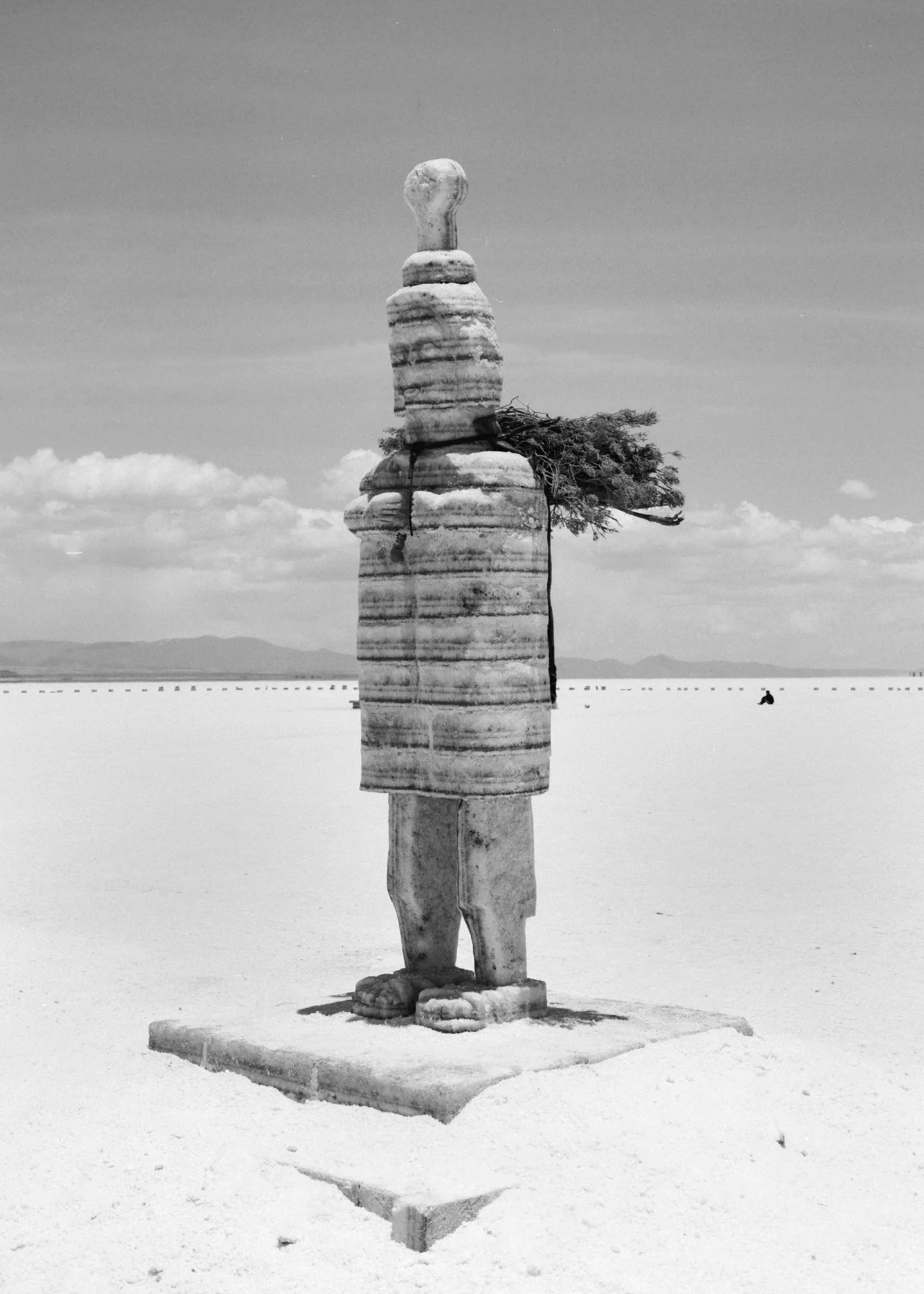 Black and white photo of a stone sculpture of a standing figure with a rounded head, set in a flat, open landscape with mountains in the distance and a single tree growing from the figure's midsection.