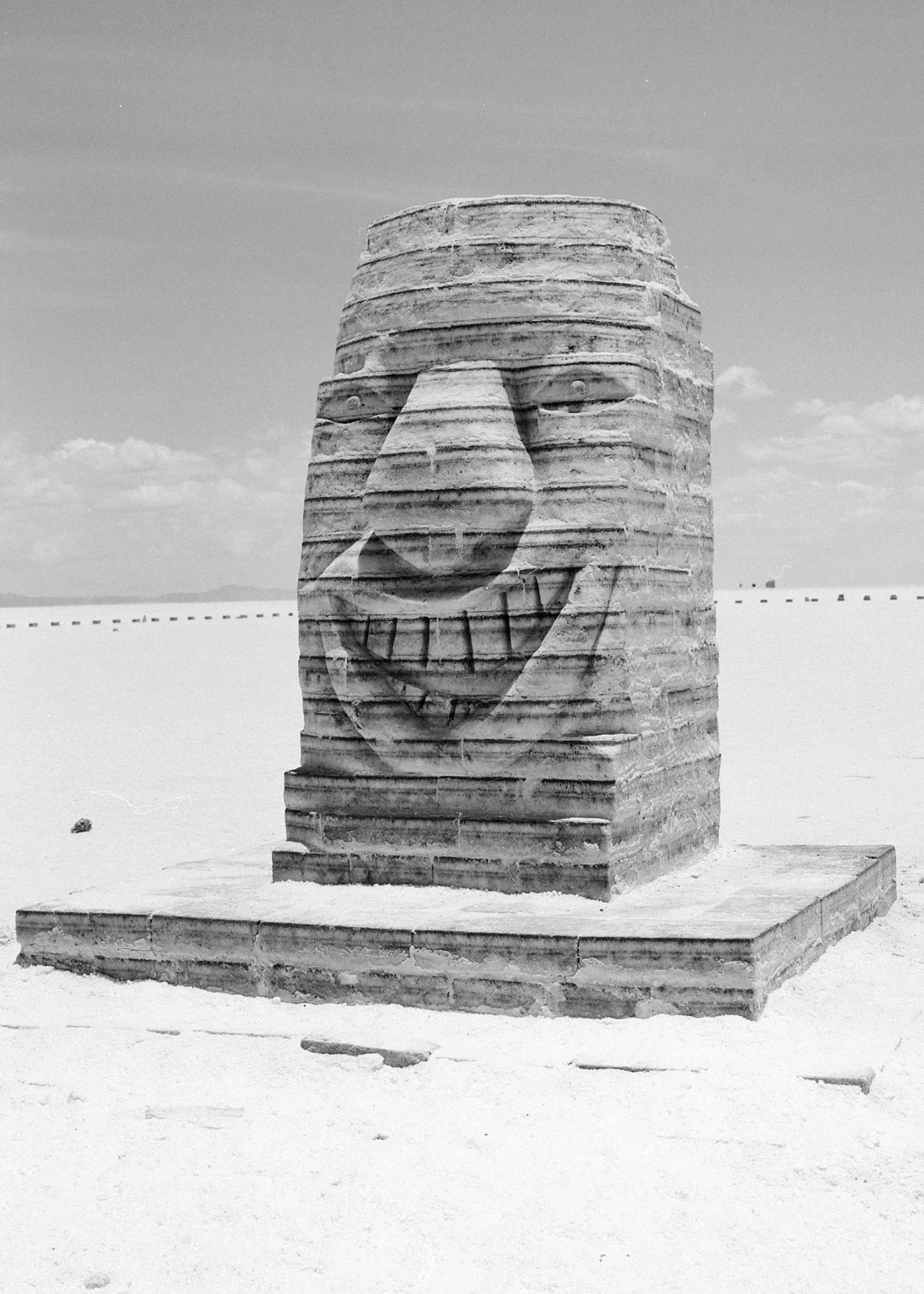 A large sculpture of a smiling face with a prominent nose and mouth, made of stacked flat stones or blocks, in a flat, open desert landscape with a clear sky.