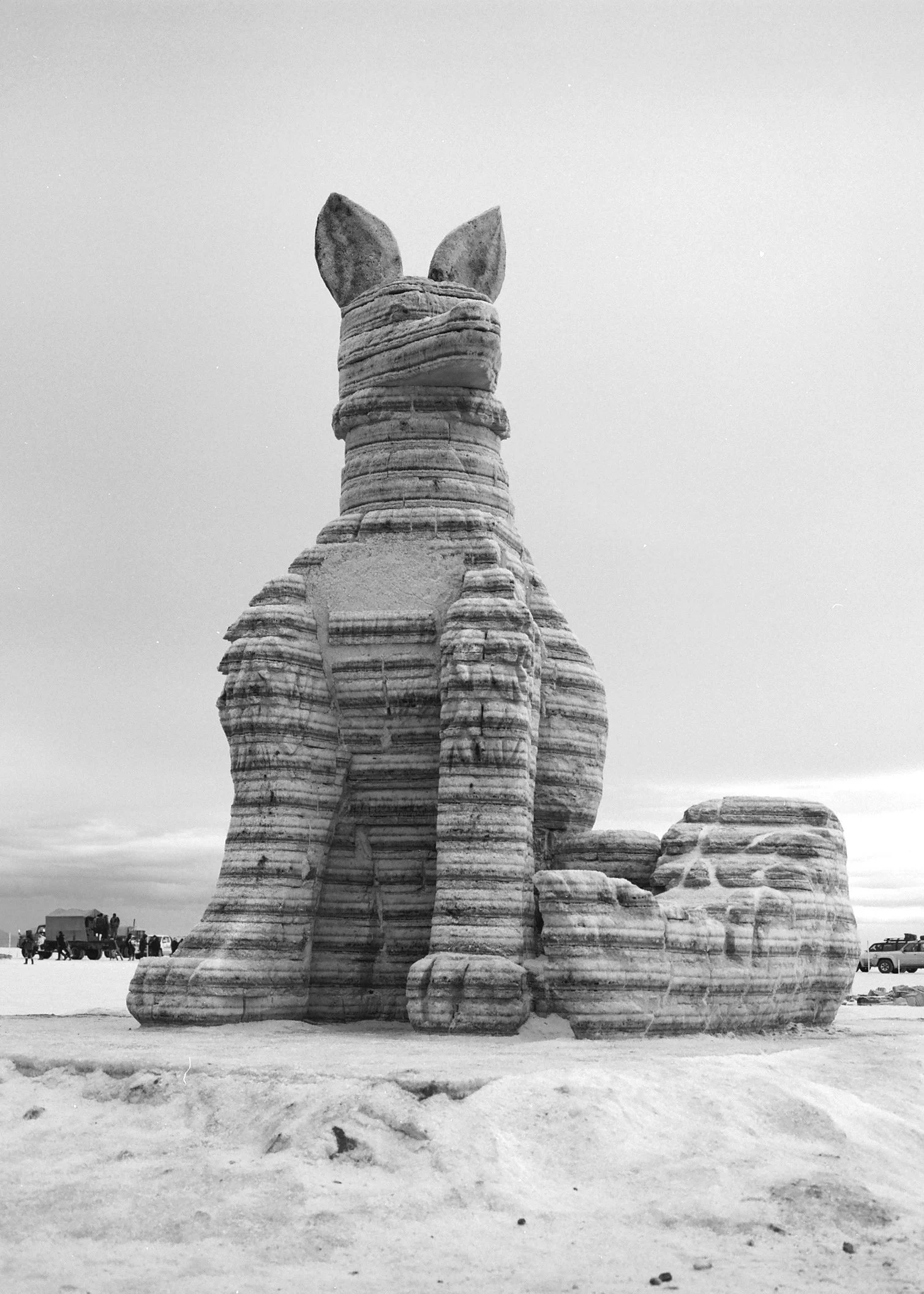 A large sculpture of a dog made from layered stone in a desert landscape, with people and vehicles visible in the background.