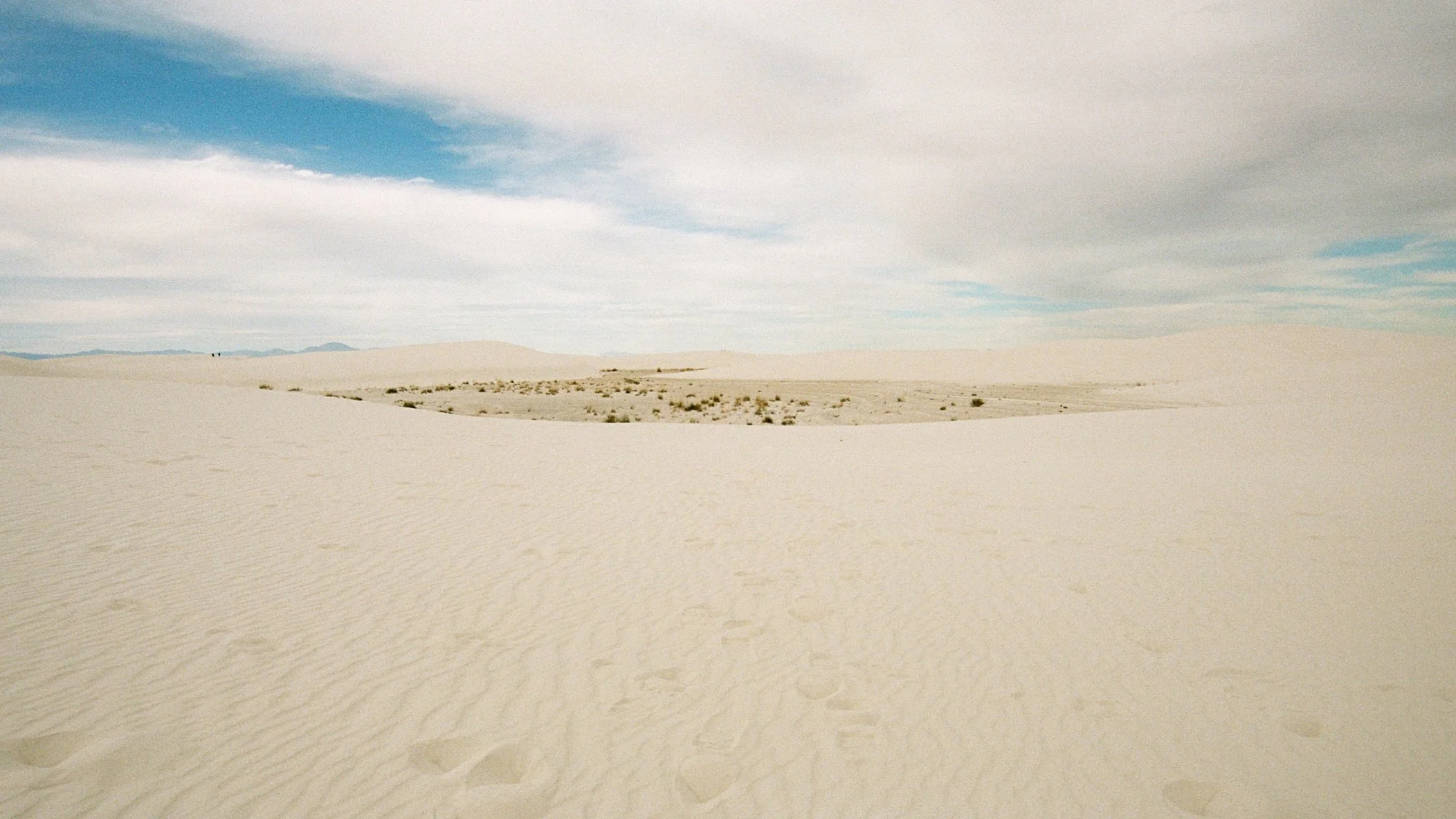 Expansive desert with rippled sand dunes, sparse vegetation, and a partly cloudy sky.