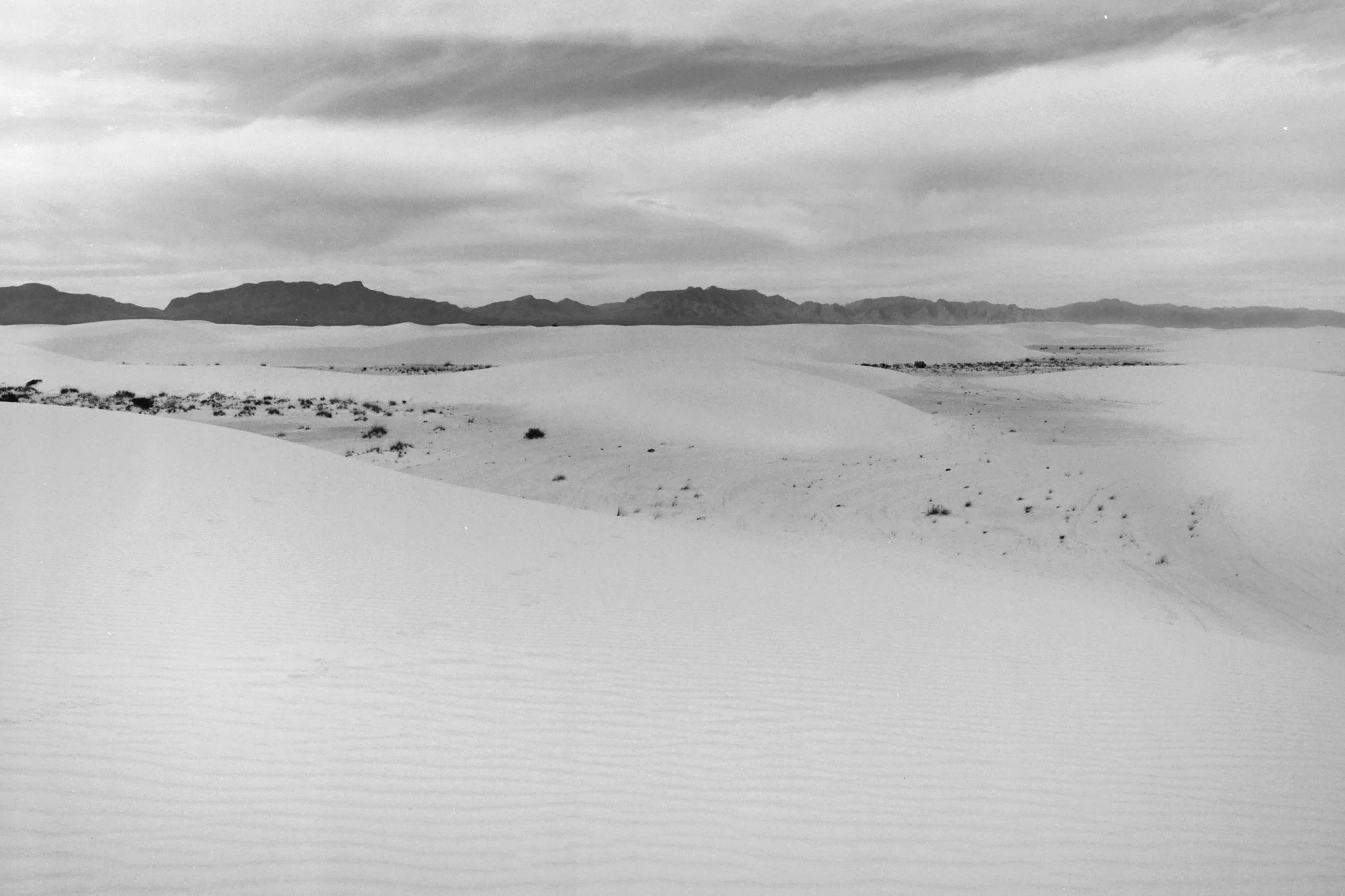 Black and white photo of sand dunes stretching across the landscape with mountains in the distance and a cloudy sky above.