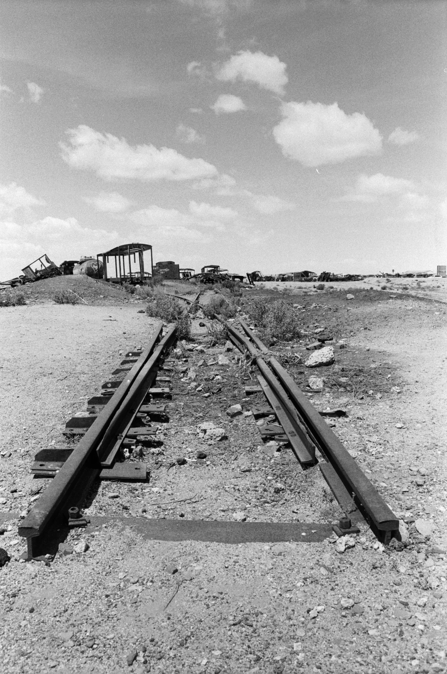 A black and white photo of abandoned train tracks in a desert landscape with rusted train cars stacked in the background, under a partly cloudy sky.