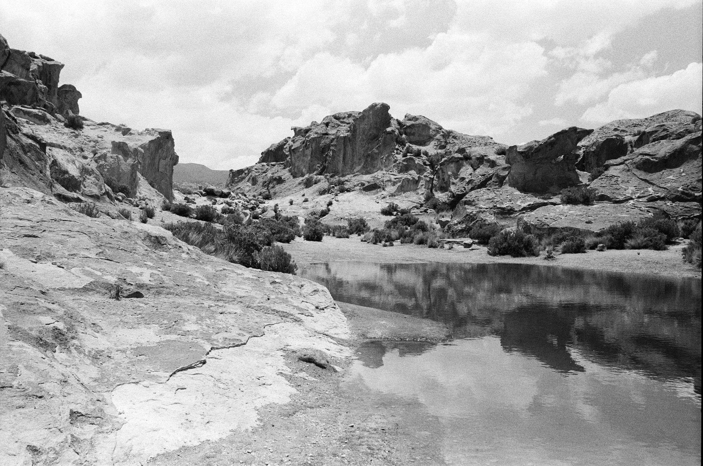 Black and white photograph of a rugged landscape with rocky cliffs, a calm river, and sparse desert vegetation under a partly cloudy sky.