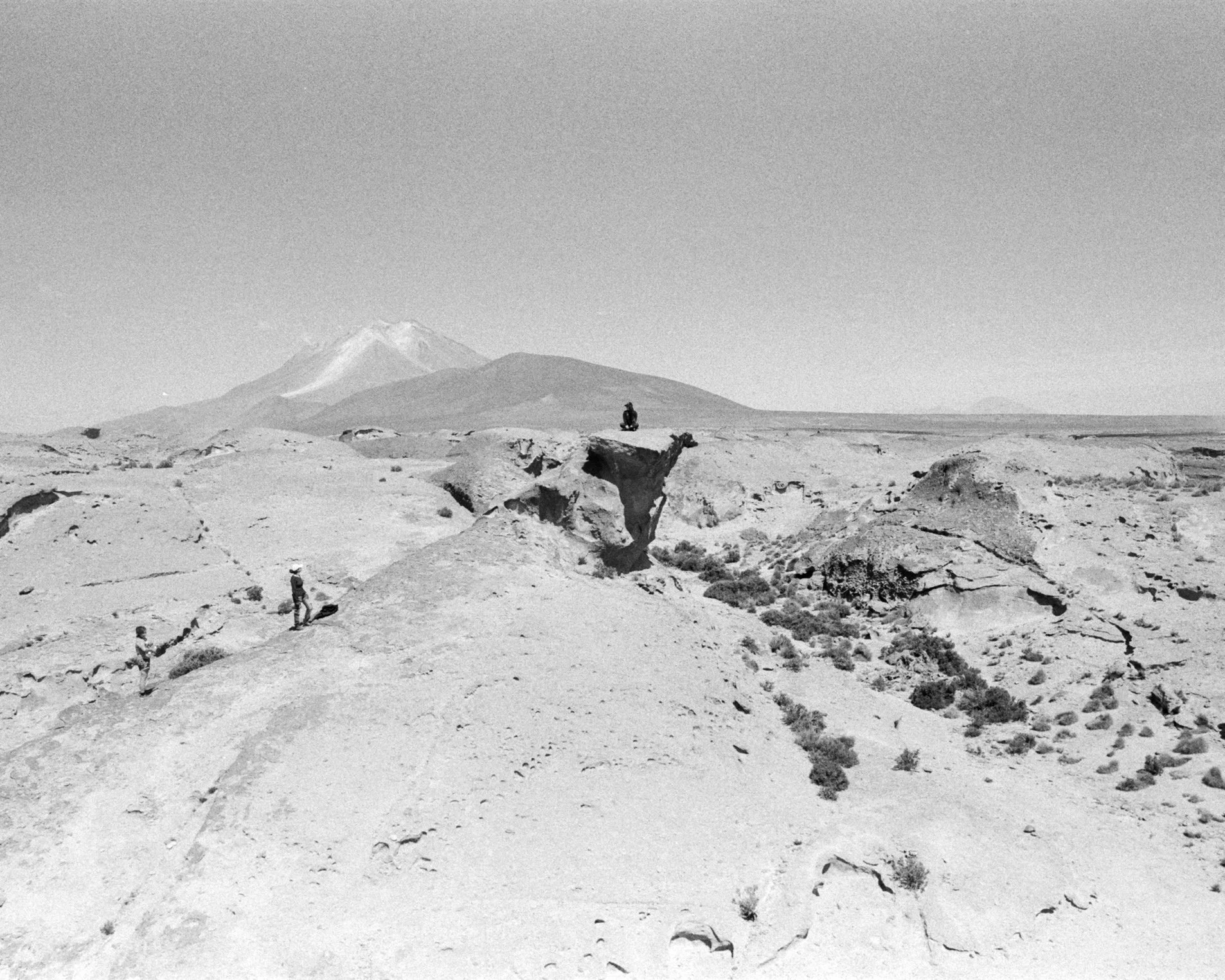 A black and white photo shows a desert landscape with three hikers walking along a dirt path. In the background, there is a mountain with snow on its peak.