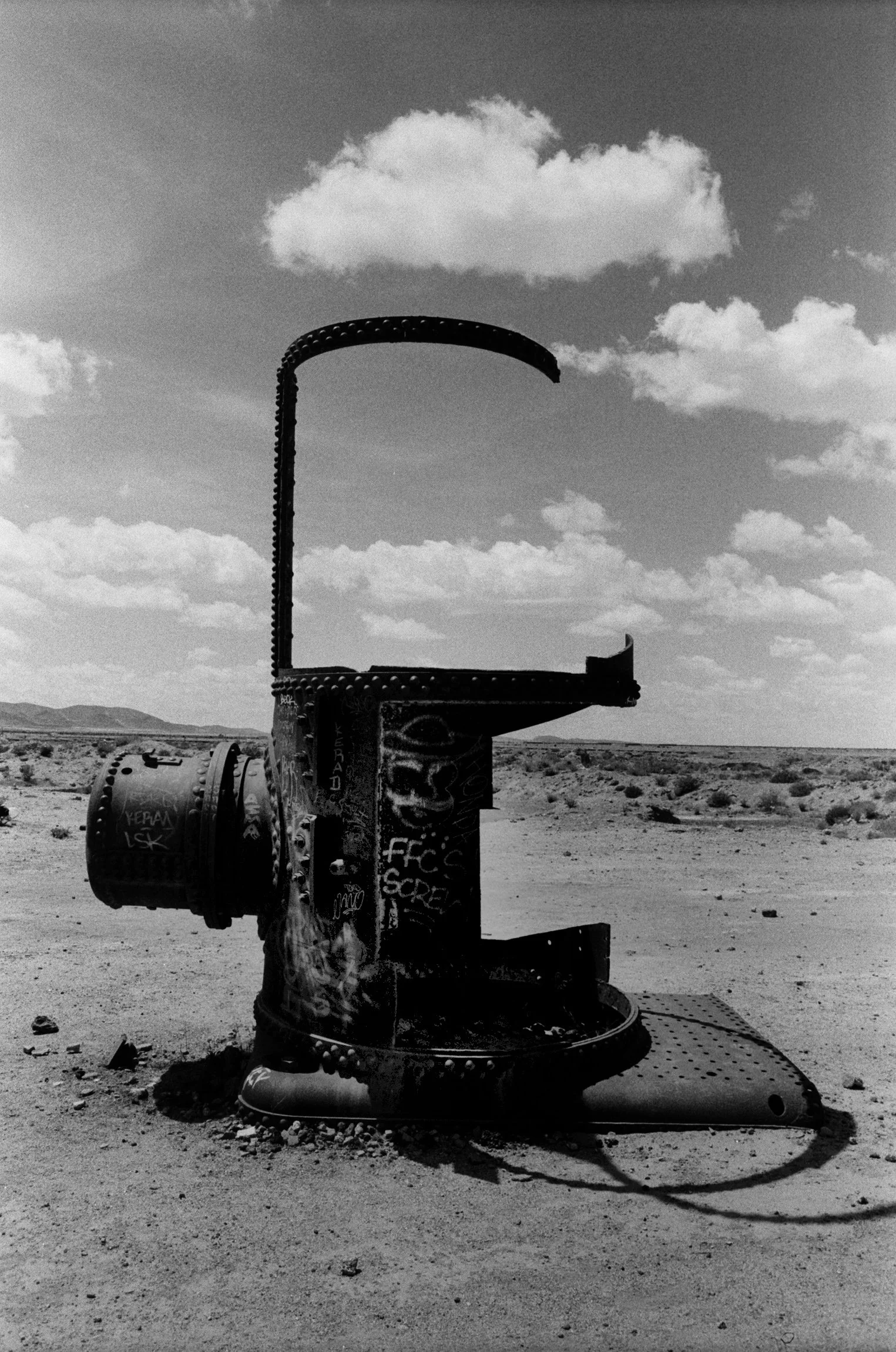 A rusty, graffiti-covered, partial metal structure in a desert landscape under a partly cloudy sky.